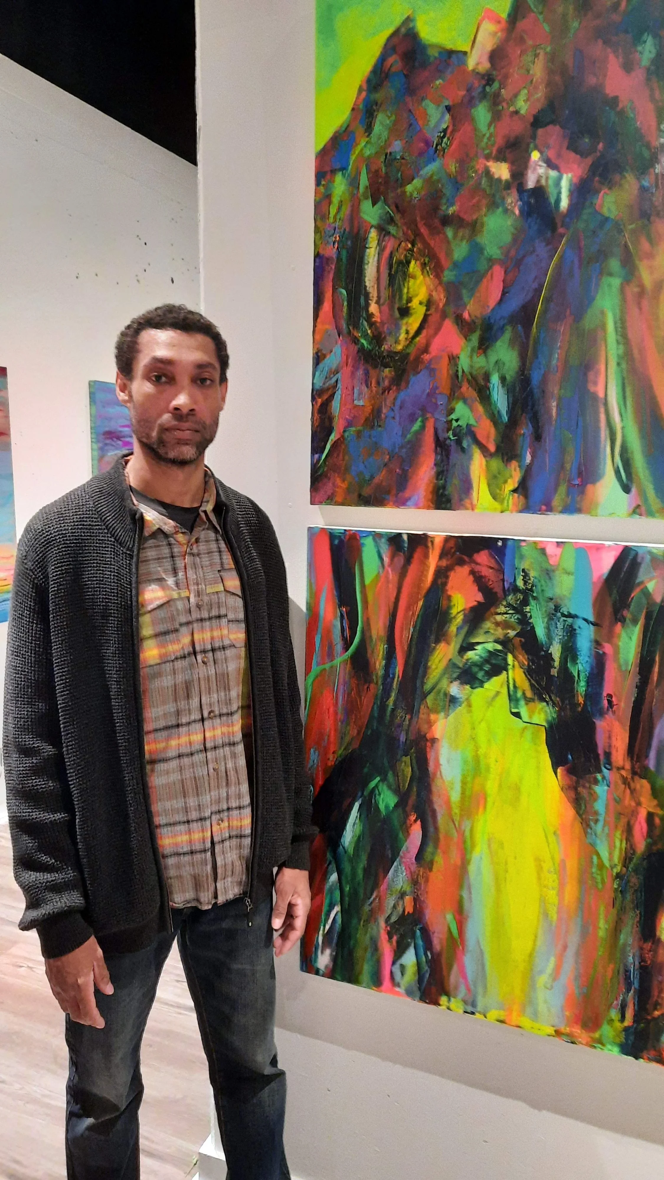 A man standing next to two colorful abstract paintings at an art gallery.