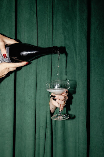 Hands pouring sparkling wine from a bottle into a coupe glass with green curtain background.