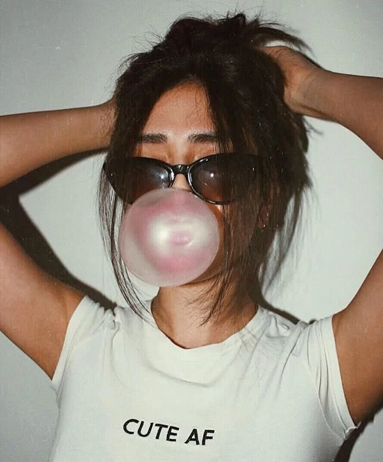 Woman in sunglasses and white "CUTE AF" t-shirt blows a pink bubble with bubblegum, hands in messyor hair.