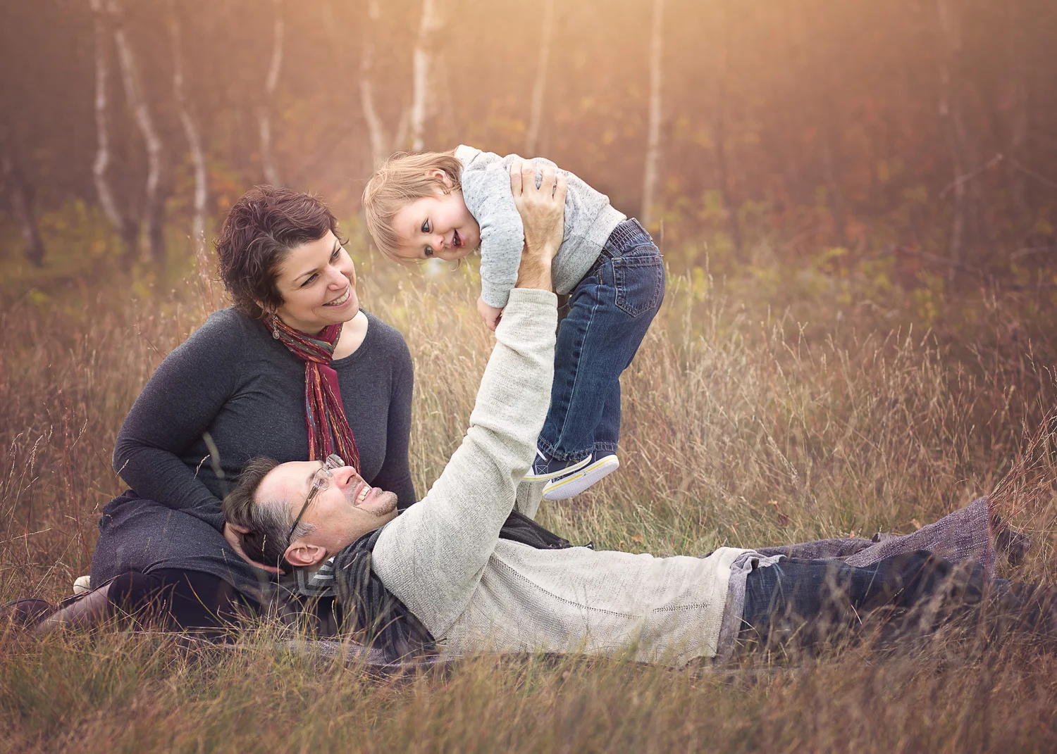 Parents lying in tall fall grass with dad holding up young boy in Birds Hill Park near Winnipeg.
