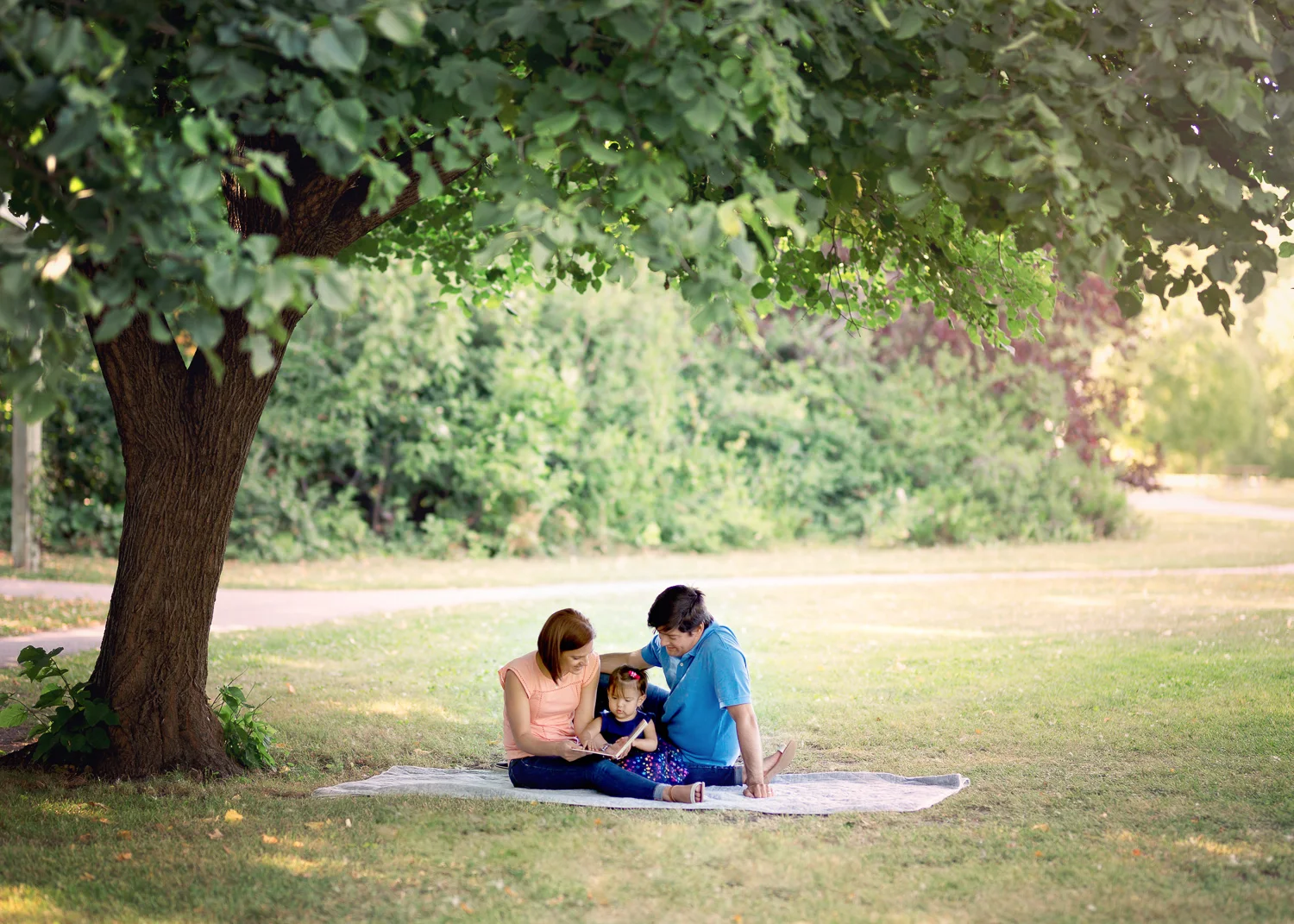 Young family of three sitting under a tree reading a book on a summer afternoon in Winnipeg.