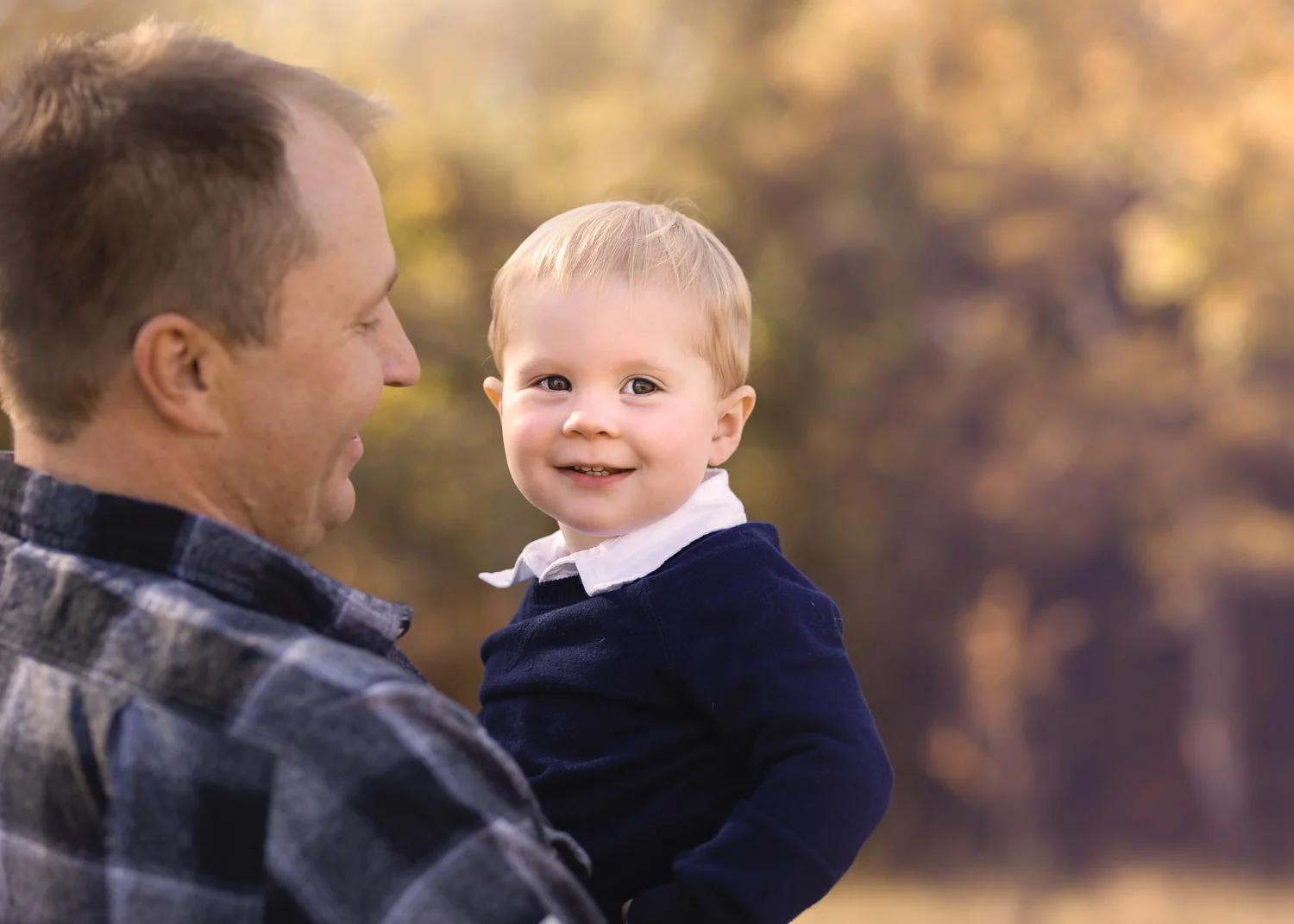 Handsome one year old boy smiling in dads arms during fall family photoshoot in Birds Hill Park near Winnipeg.