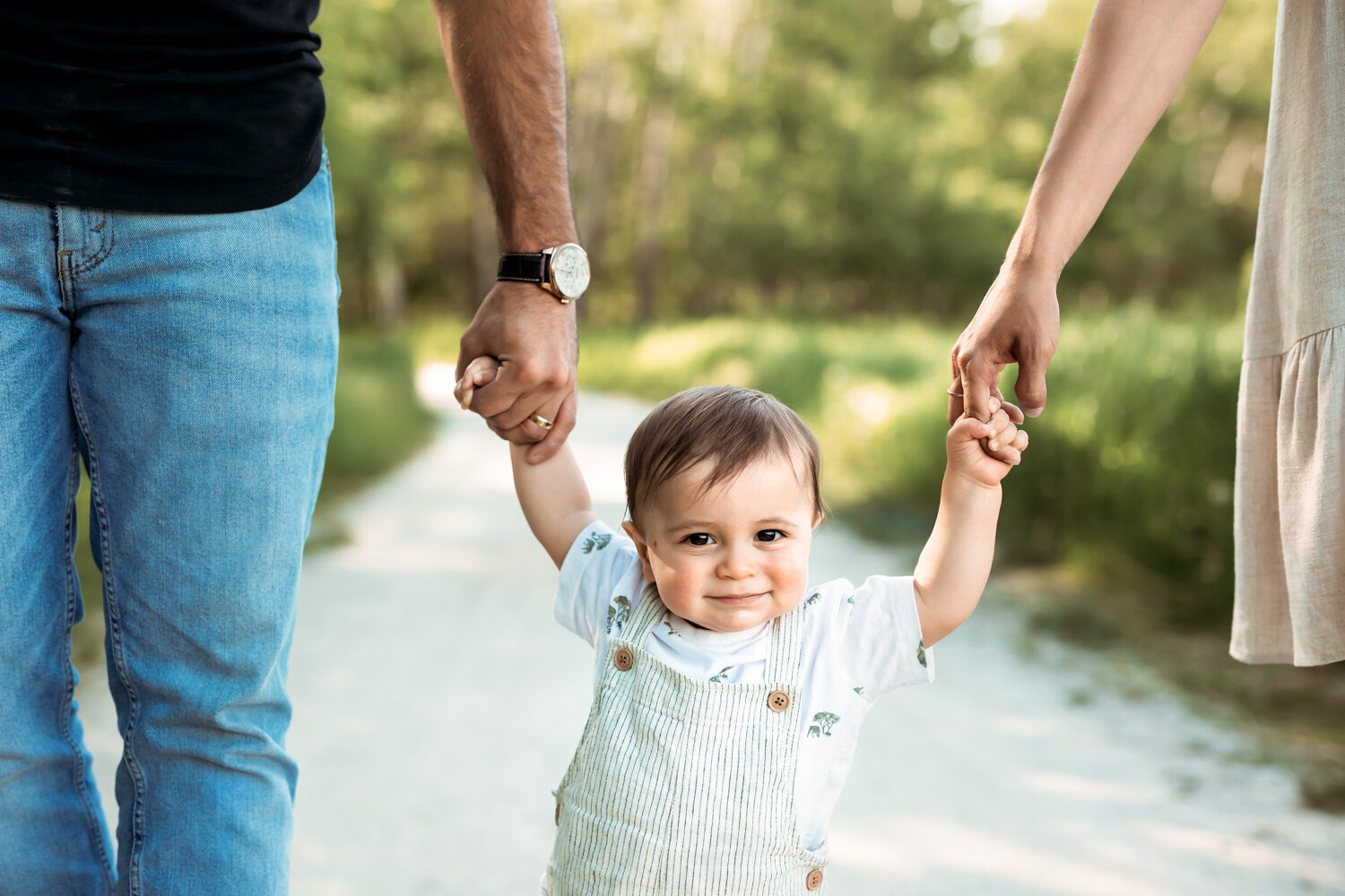  One year old boy in overalls holding mom and dads hands smiling and walking in park in Winnipeg 