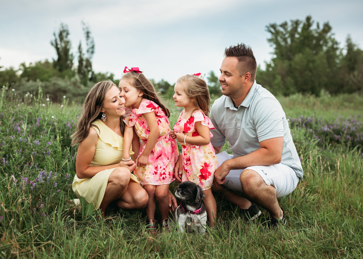  young daughter giving mom a kiss while dad and sister watch during photoshoot in the grass and flowers near WInnipeg 