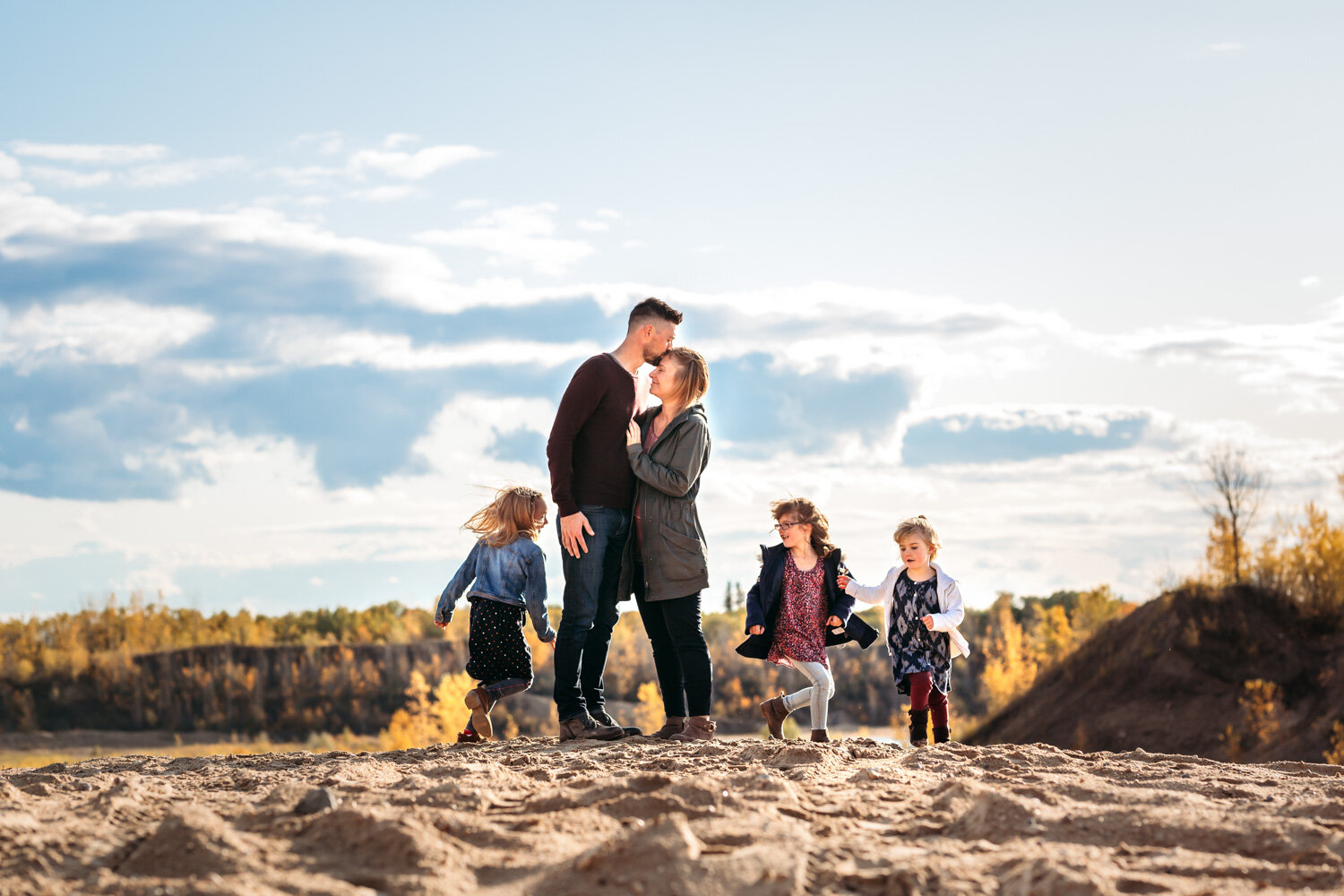  Daughters running around parents embracing during fall family photoshoot sand dunes near Winnipeg 