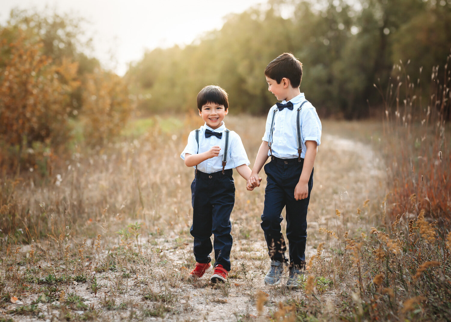  Two young brothers in cute bowties walking hand in hand and smiling with sun shining behind them 