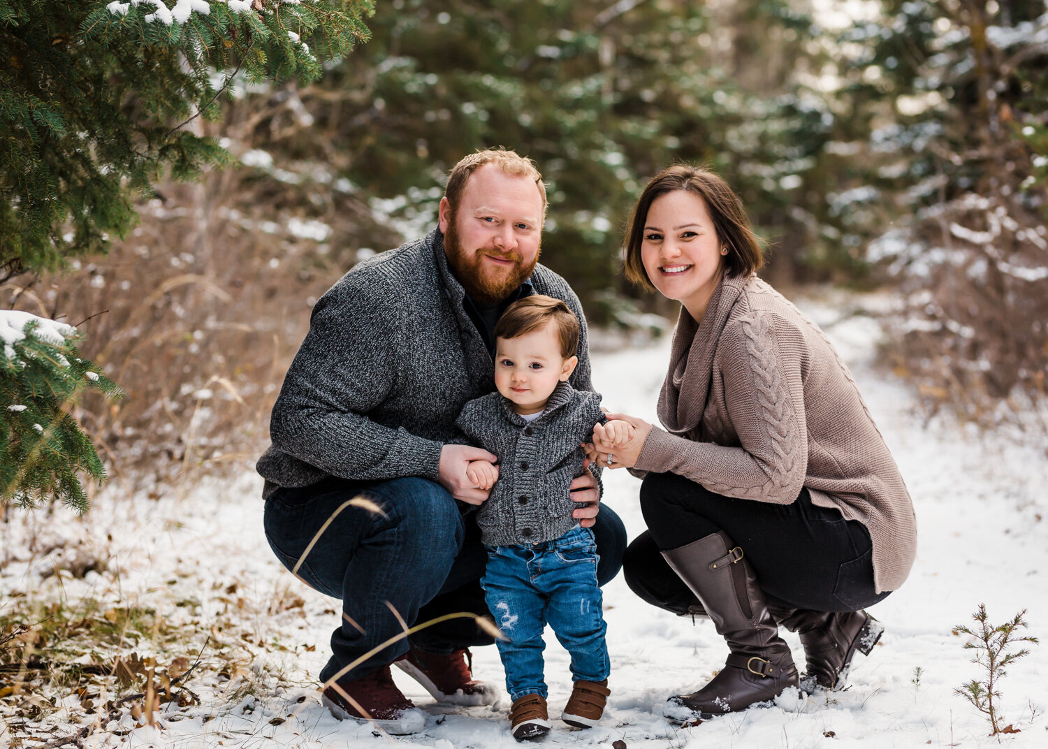  Cute one year old boy with parents crouched down beside him in snowy pines at Birds Hill park near Winnipeg 