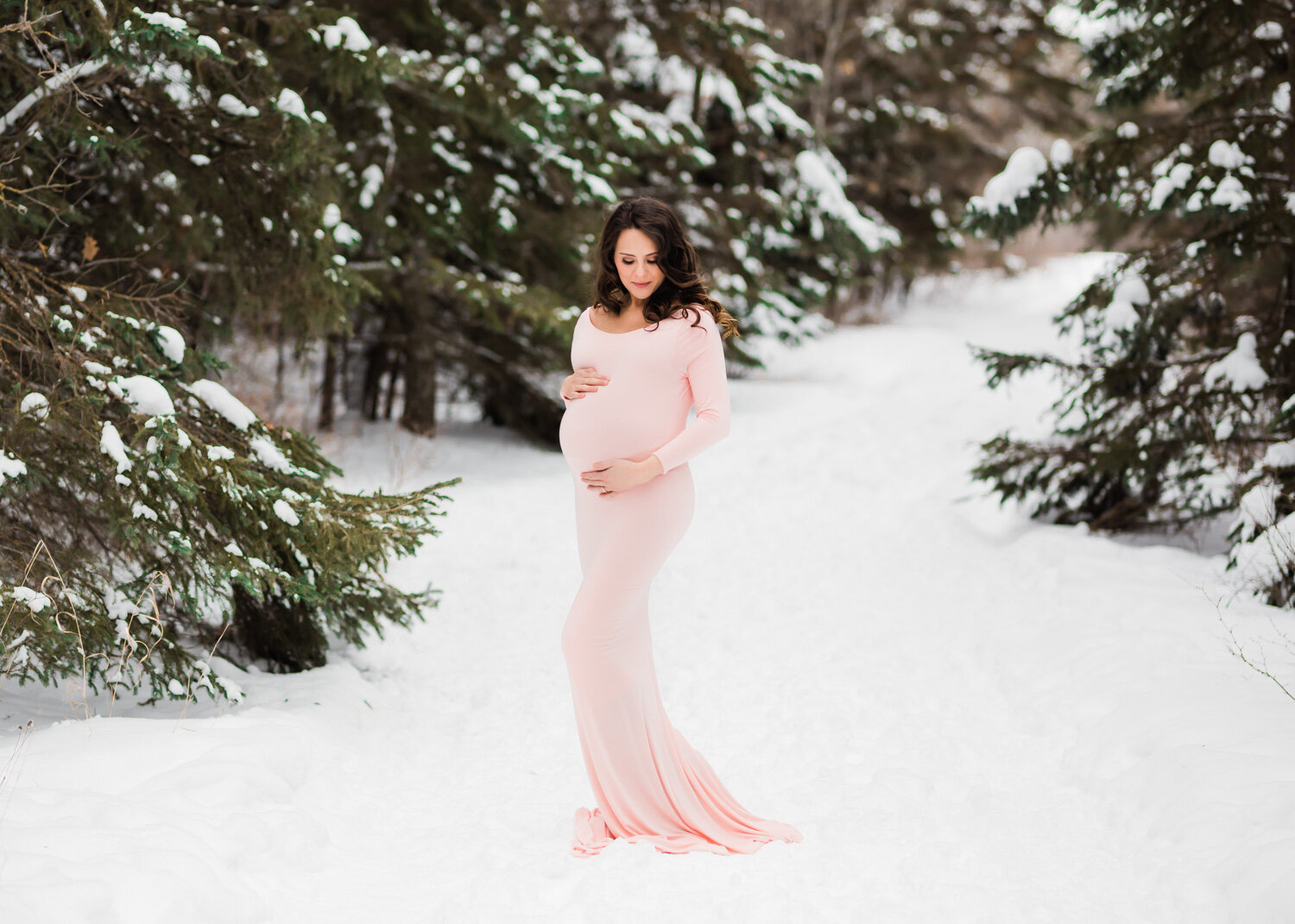  Radiant pregant mother holding belly in blush maternity gown in forest of snowy Winter Evergreens near Winnipeg 