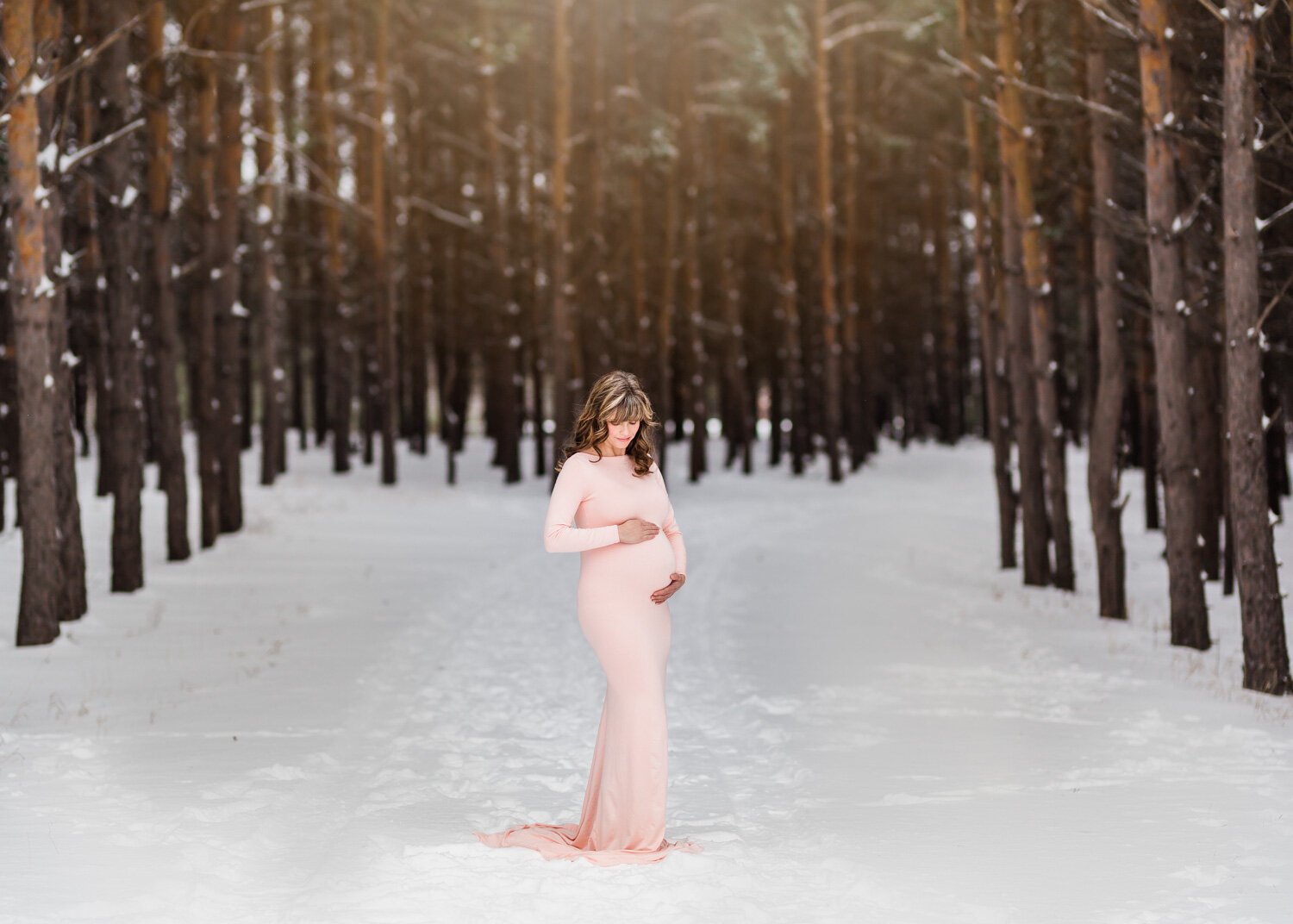  Woman looking down at pregnant belly in gorgeous blush pink maternity dress in tall pines near Winnipeg 