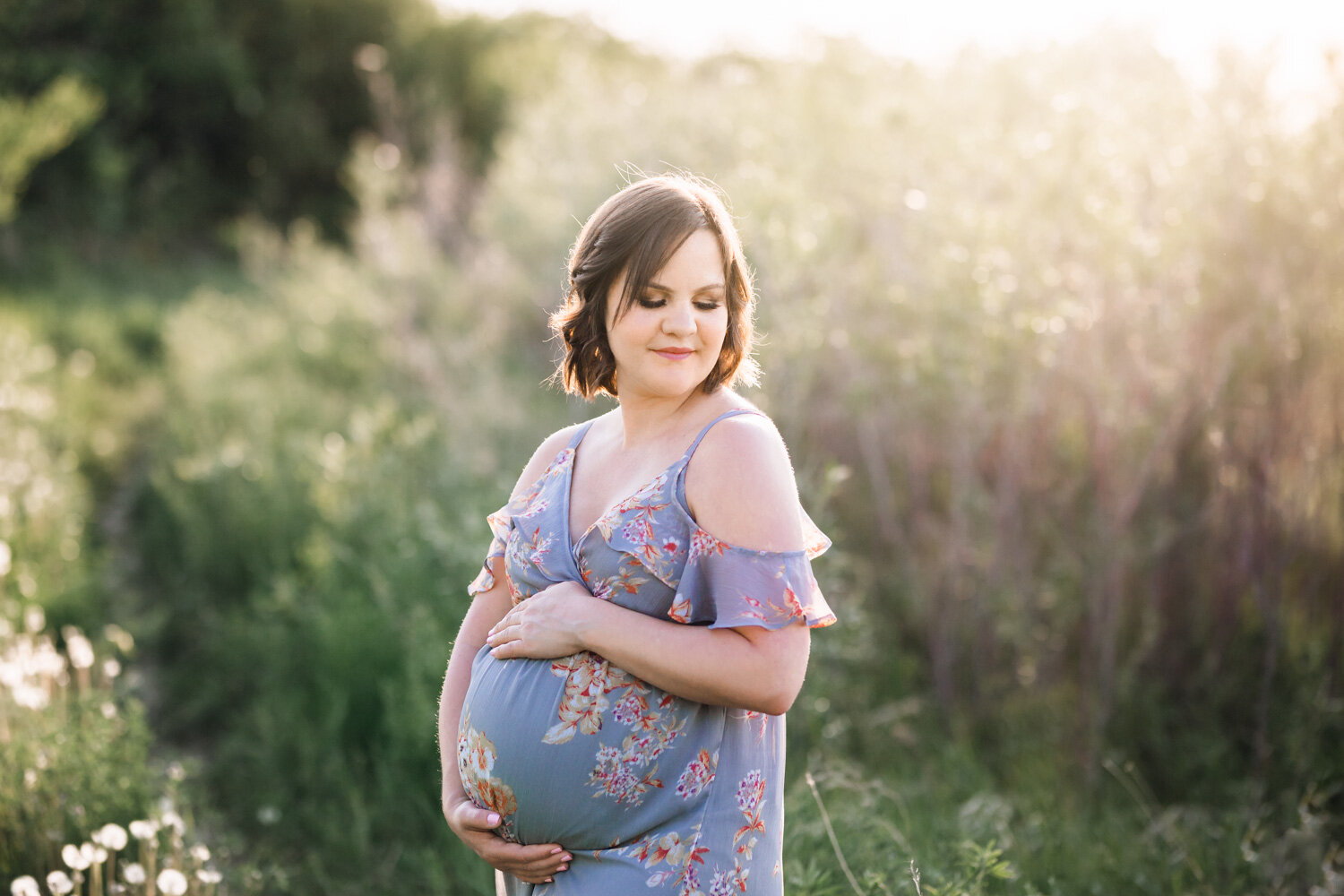  Woman casually looking over shoulder holding pregnant belly outside in spring field for maternity photos near Winnipeg 