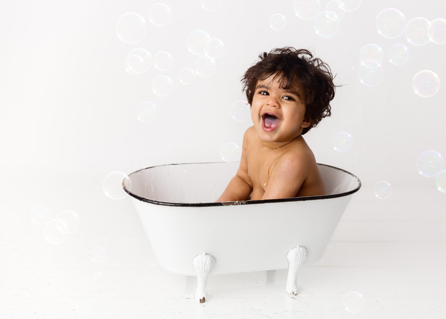  Laughing jovial boy in white antique tub with floating bubbles after cake smash photography session in Winnipeg 