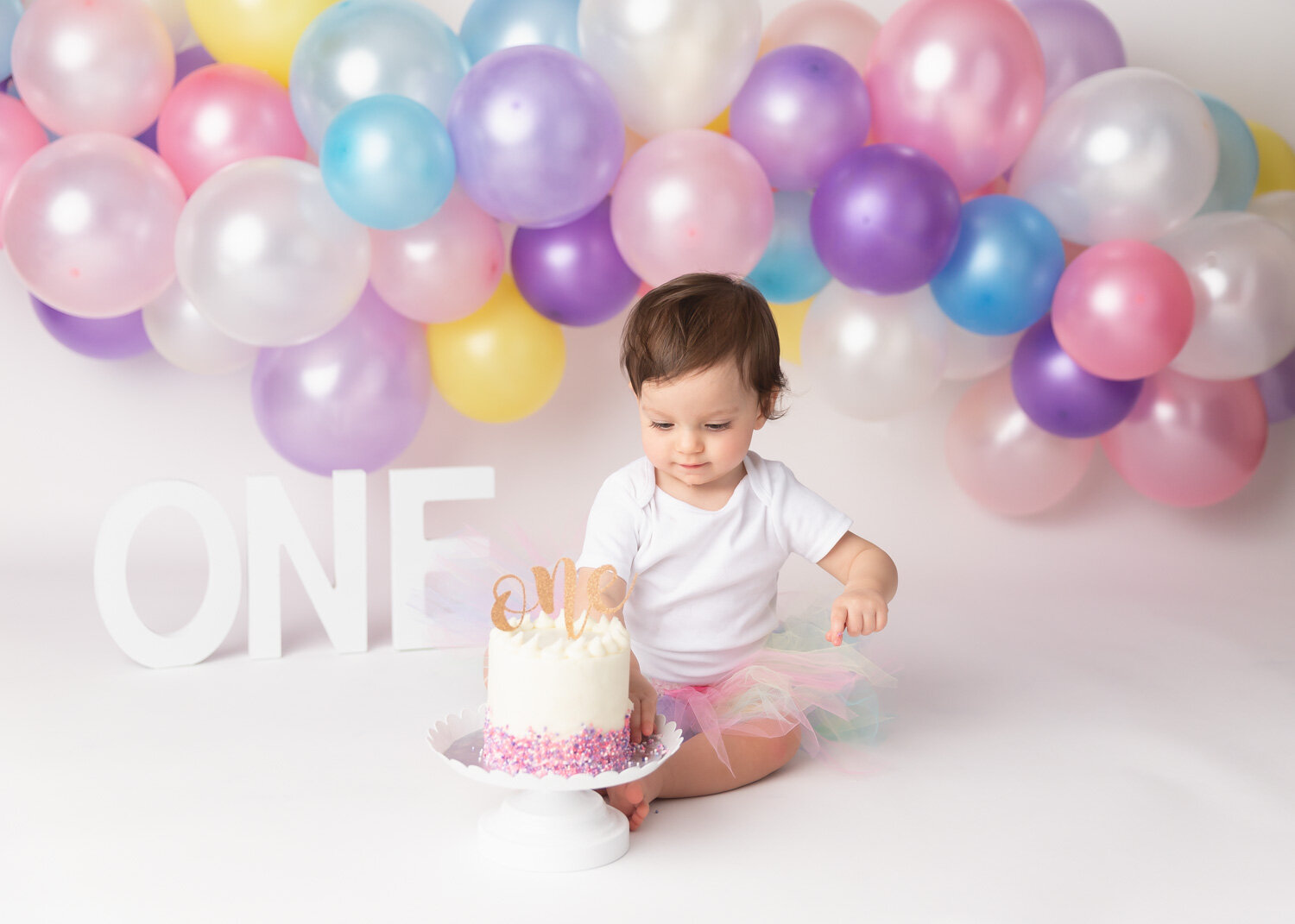  Girl starting to dig into cake smash with pastel rainbow balloon garland in Winnipeg photography studio 