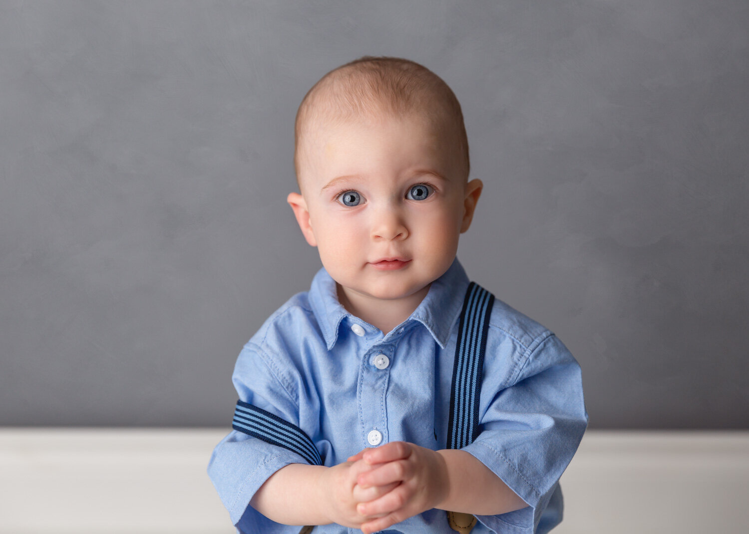  Big blue eyed one year old boy with blue suspenders sits with hands clasped for child portait session in Winnipeg 