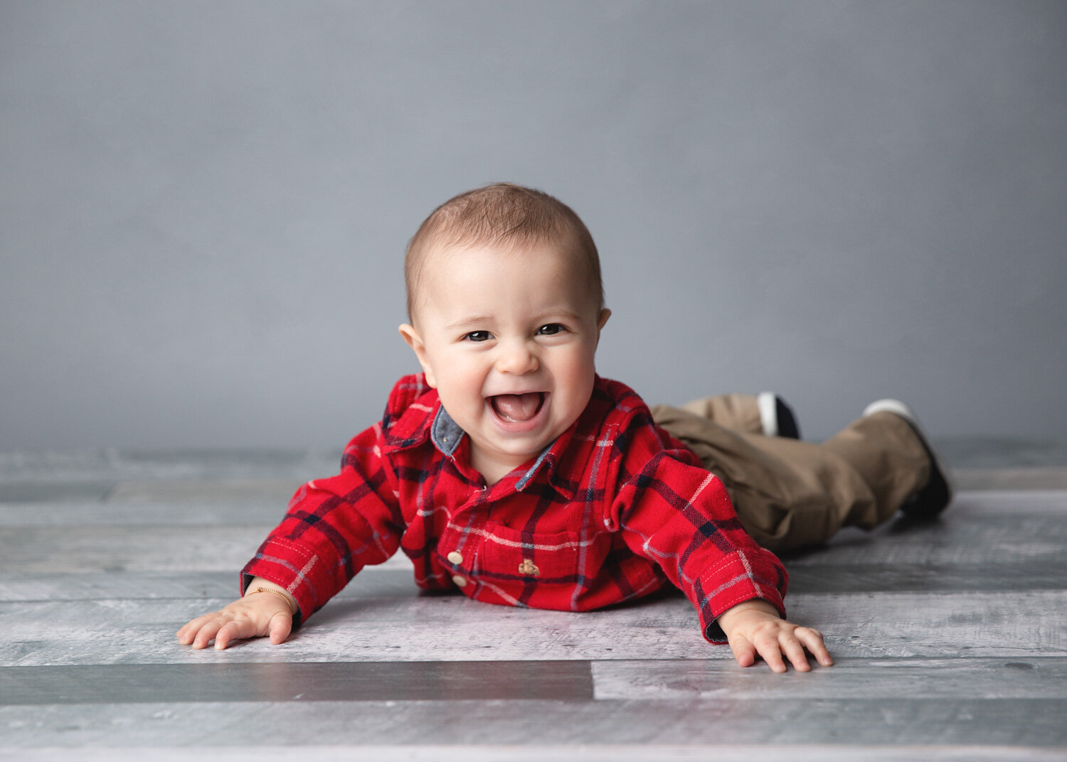  Happy boy laughing in red plaid shirt lying on tummy on grey wood floor during sitter child milestone session in Winnipeg 