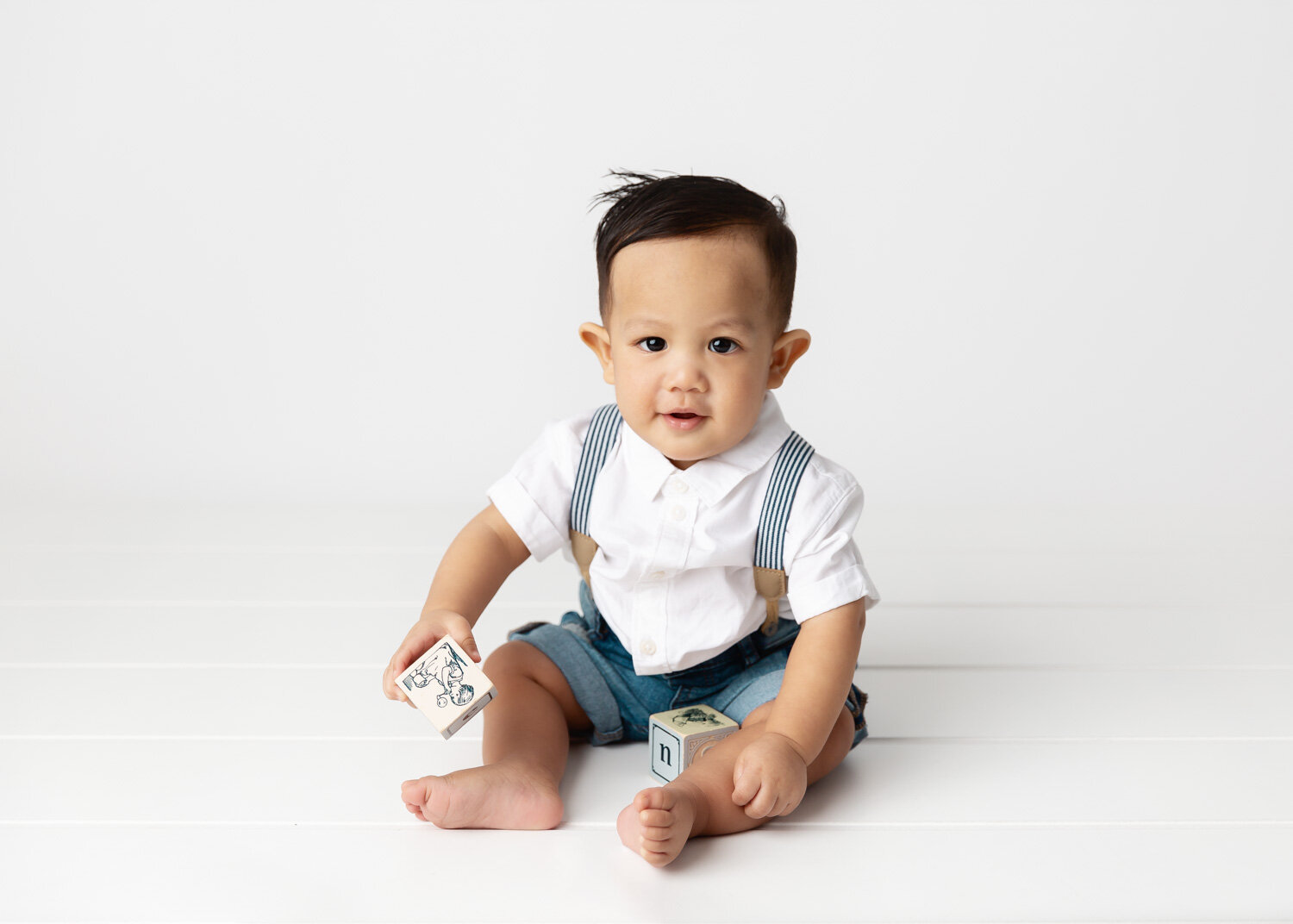  Handsome baby boy in jean shorts and suspenders playing with blocks during child photography session in Winnipeg 