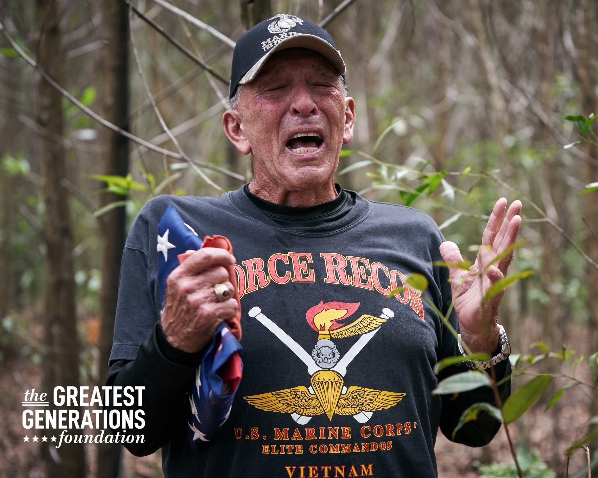 Elderly man in a forest, wearing a U.S. Marine Corps Force Recon shirt, holding an American flag. He's emotional and the image is associated with "The Greatest Generations Foundation."