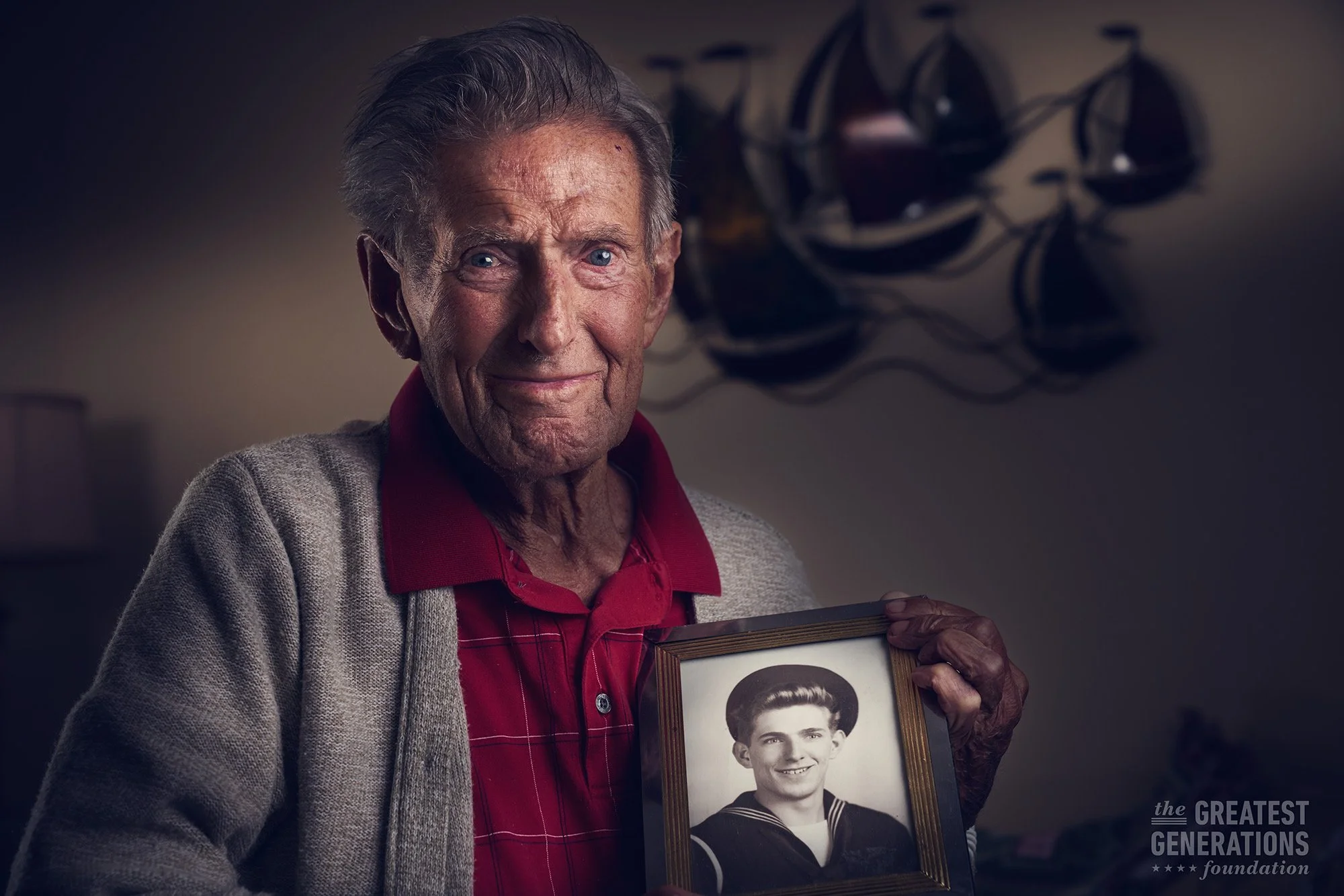 An elderly man holding a framed black-and-white photo of a young man in a sailor uniform, standing in a warmly lit room with wall decor in the background.
