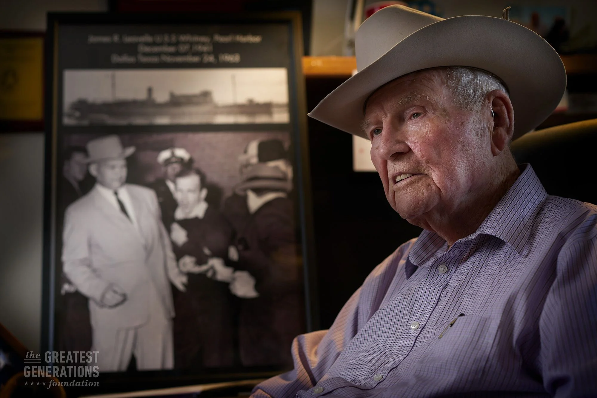 An elderly man wearing a cowboy hat and a checkered shirt sitting indoors, with a large black and white photo on an easel behind him that displays a historical group photo of people in vintage clothing.