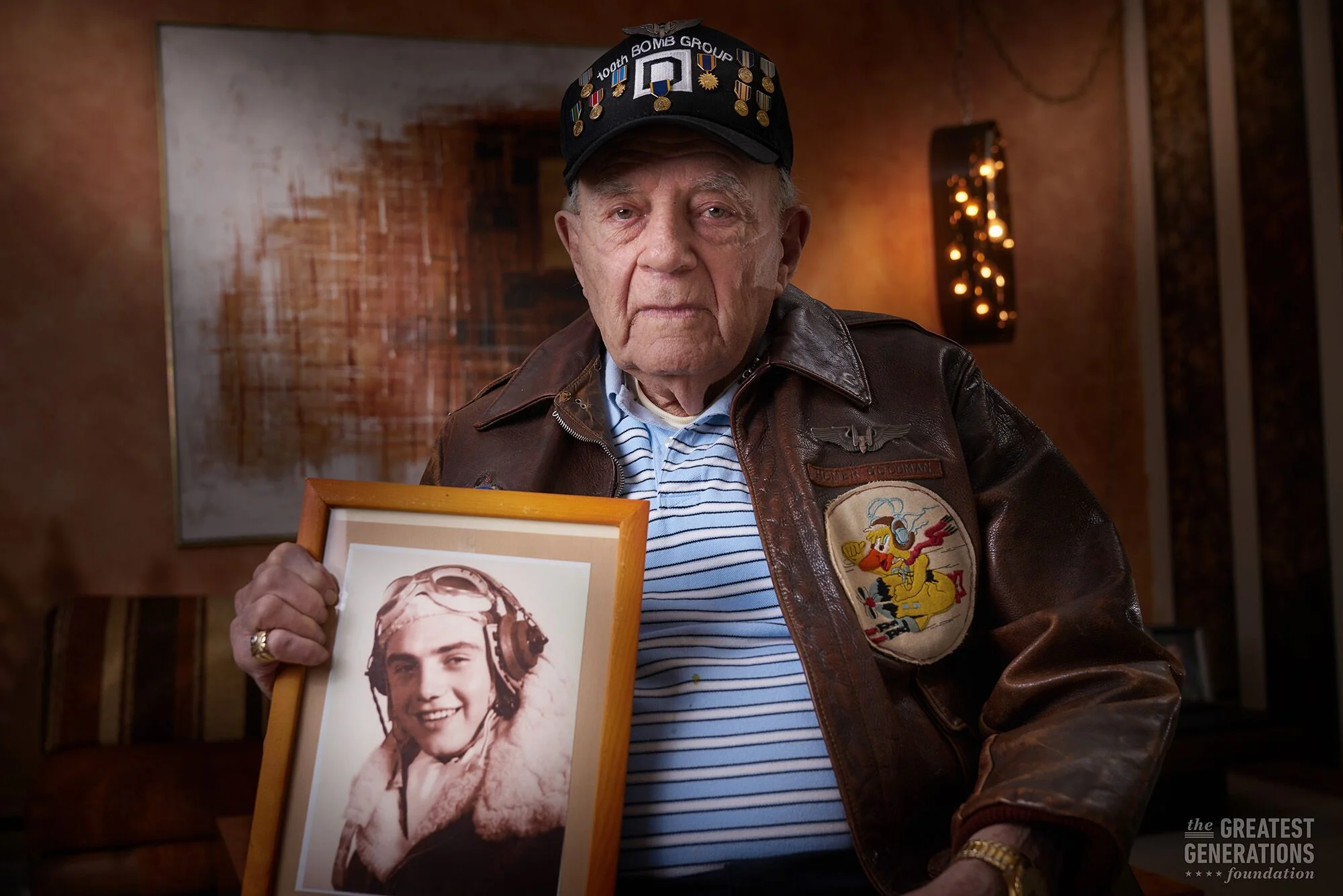 An elderly man in a leather jacket and a cap with medals, holding a framed photo of a young woman in military uniform, inside a warmly decorated room.