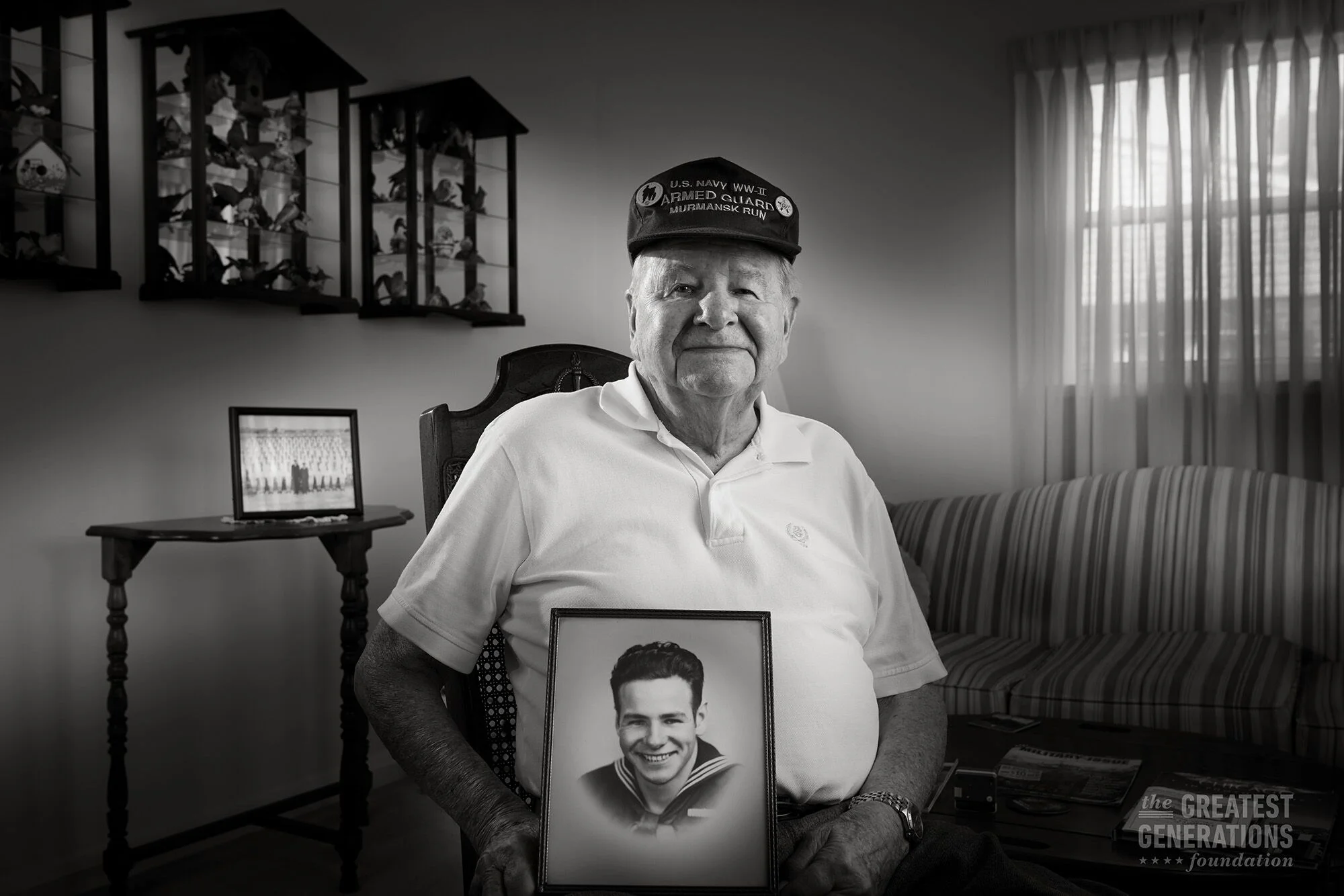 An elderly man with a hat sitting in a chair, holding a framed photograph of a young man in a naval uniform.