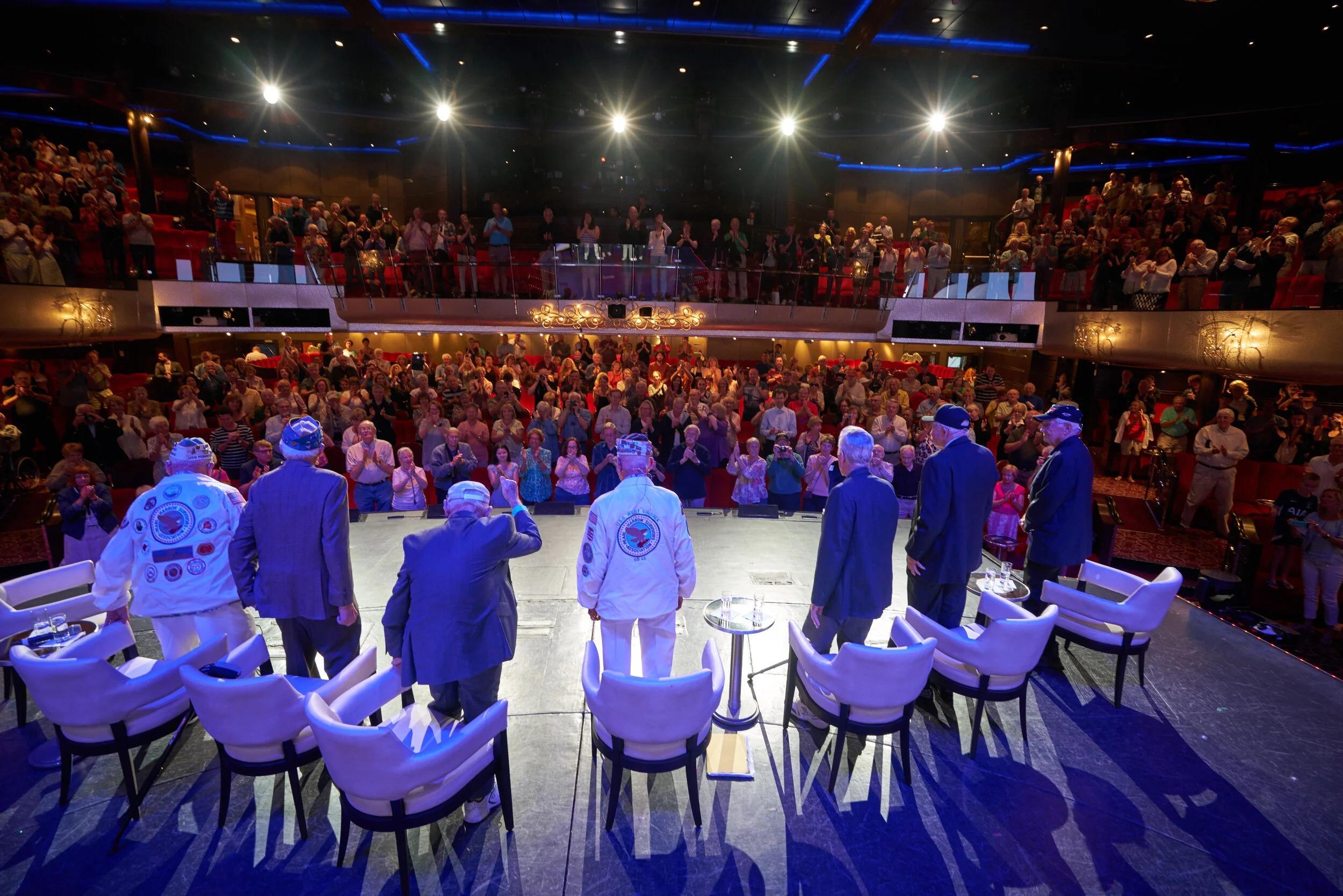 Stage with six men, some in astronaut jackets, facing an audience in a theater. Audience members are clapping, and the scene is lit with bright stage lights.