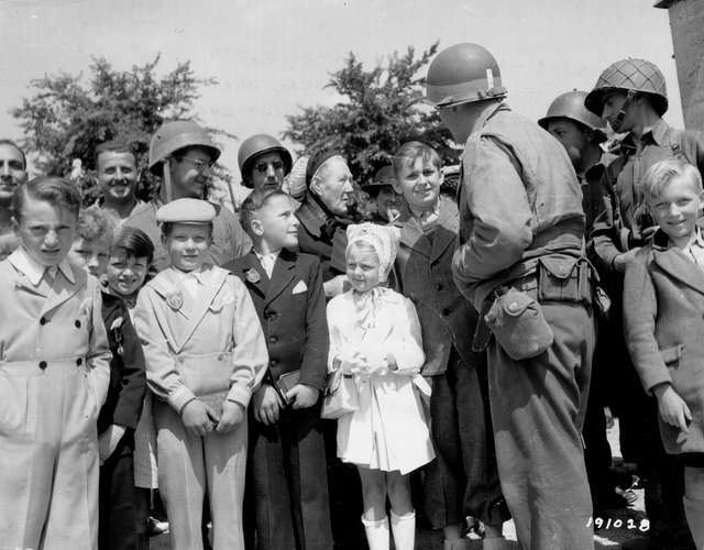 Group of smiling children in formal clothing gathered around soldiers in uniform helmets during World War II. Trees and sky in the background.