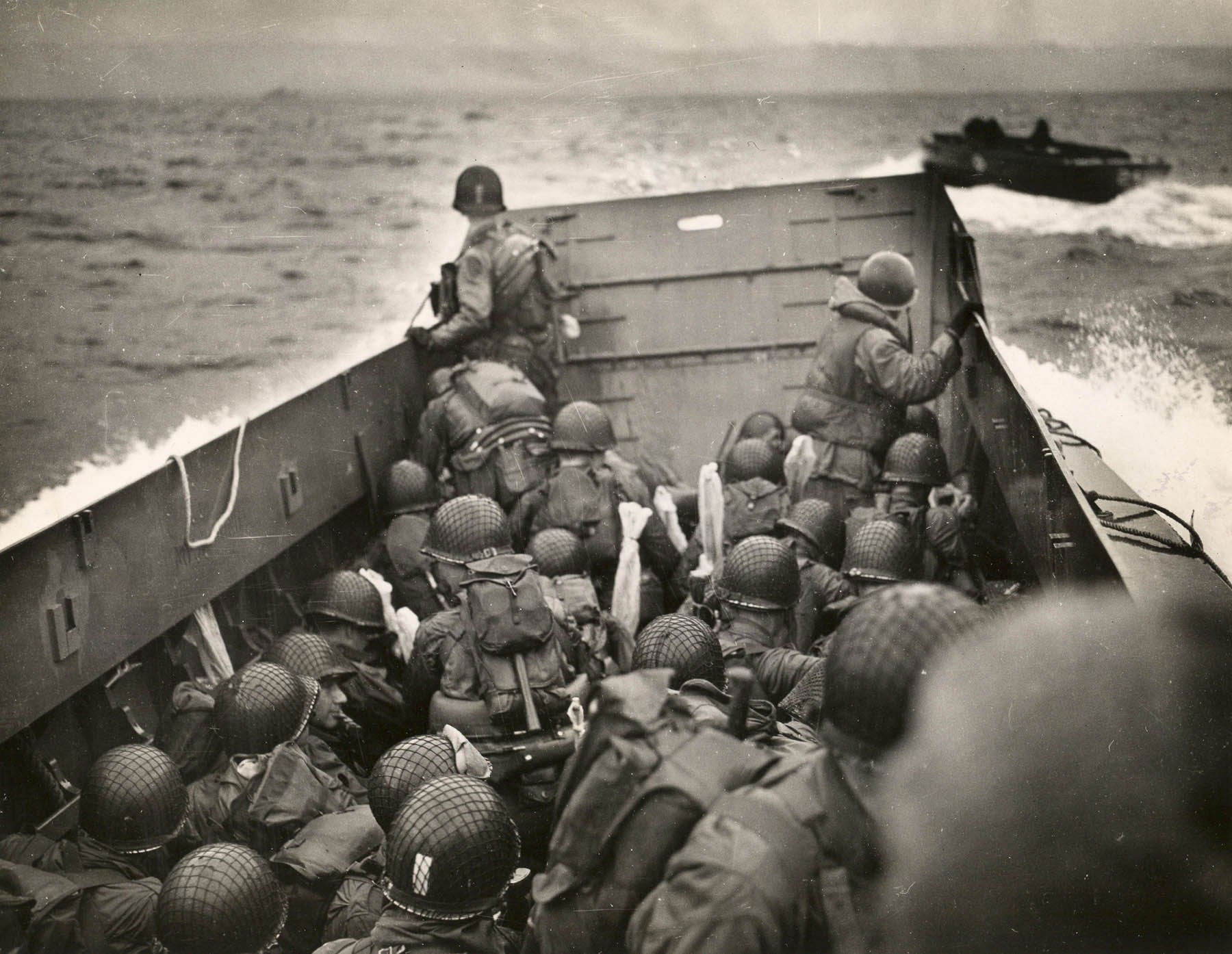 Soldiers in a landing craft during a military operation, wearing helmets and gear, approaching a beach with another boat visible in the background.