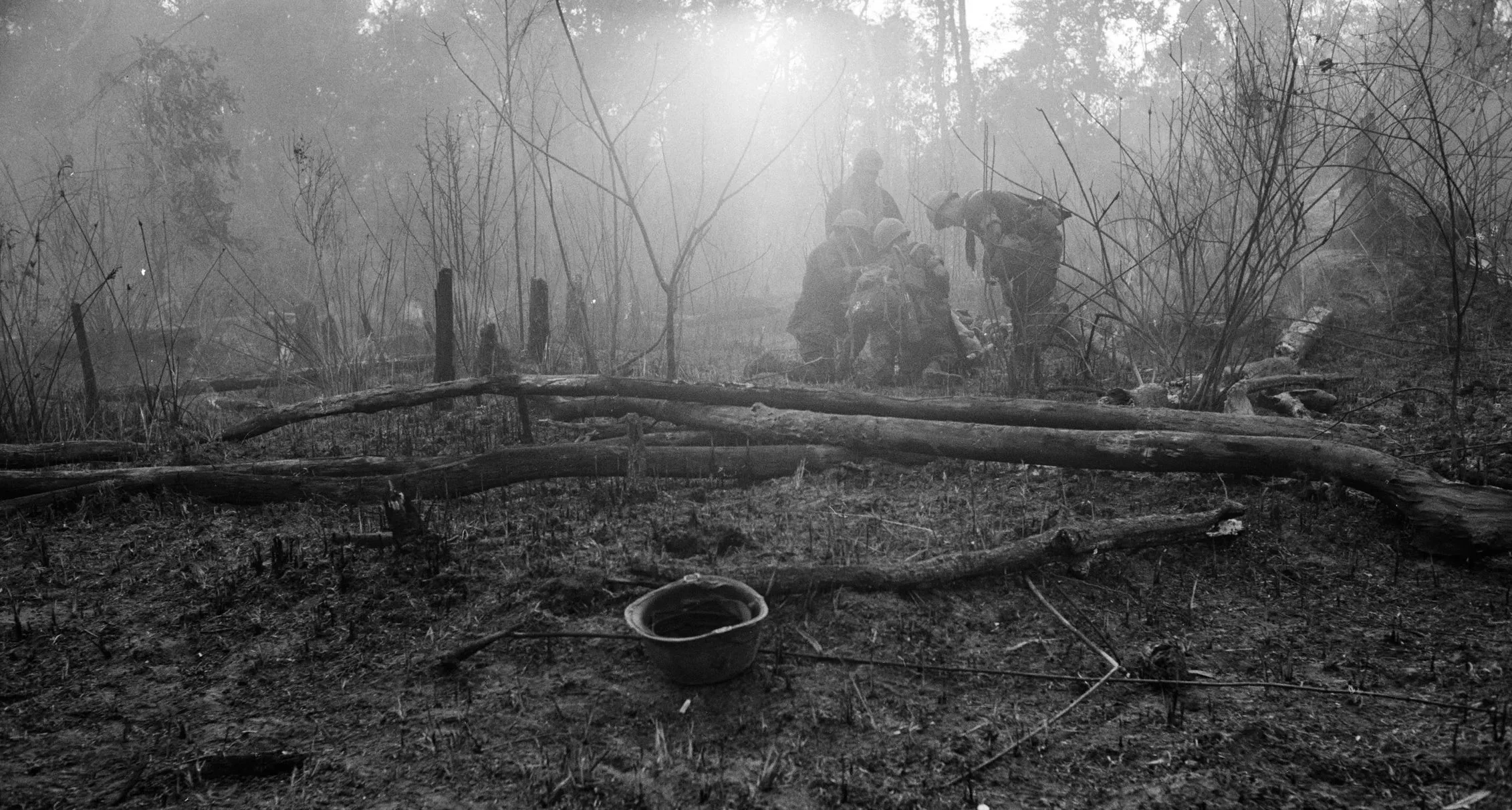 Soldiers helping each other in a smoky, damaged forest with fallen trees and debris.
