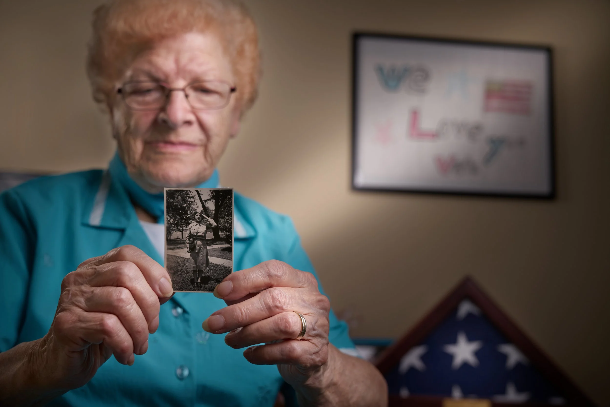 An elderly woman with glasses holding a black and white photograph of herself outdoors, with an American flag folded in a triangular display case in the foreground.