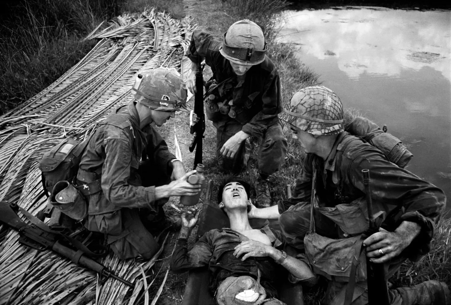 Black and white photo of soldiers rescuing a wounded person lying on the ground near a body of water.
