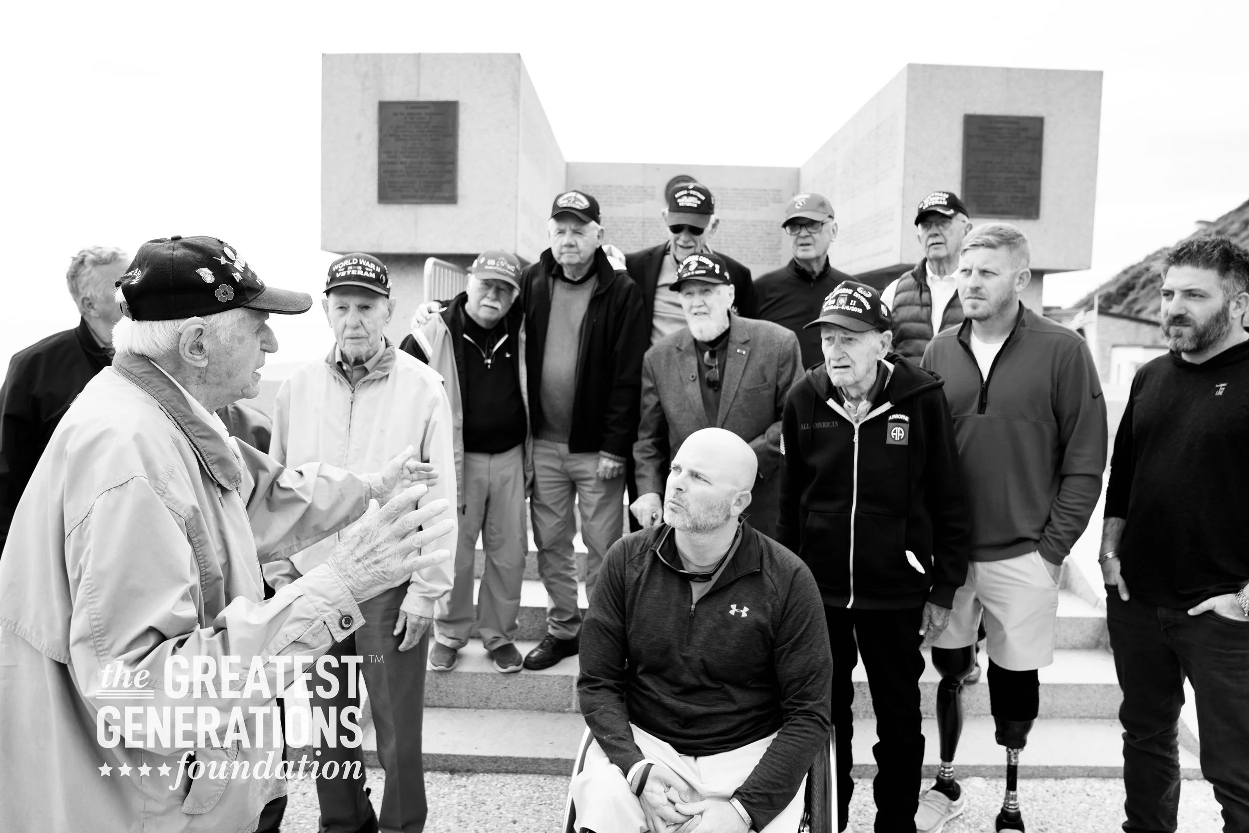 An elderly man wearing a "2nd Armored Division WWII" hat and glasses standing next to a smiling young man holding a lollipop. They appear to be at an event hosted by The Greatest Generations Foundation. The older man has his arm around the younger man's shoulder.