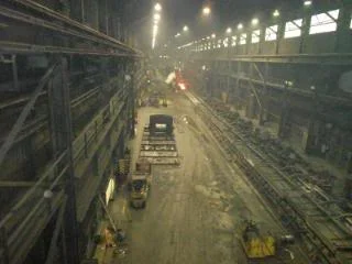 "Crane view midway of the Hot Strip Mill looking south towards the furnace area at the end of the mill. The yellow apparatus was used to change finish mill work rolls. Past the tow motor is the large backup finish mill rolls. Farther down you can see the Hot strip coming out of the 4 high Roughing mill. Past that is a Two High 'Scale' breaker. It reduces the 9 inch thick hot strip to 7 inches in one pass while extreme water pressure blasts the scale off of the strip. At the end of the building is the one Walking Beam Furnace that replaced the 3 pusher type furnaces."