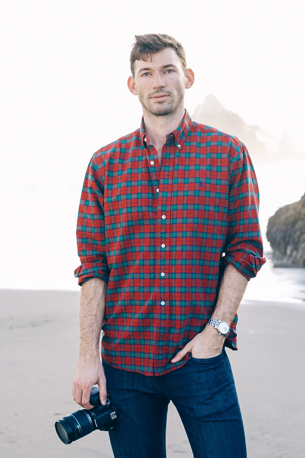 Man standing on a beach holding a camera, wearing a red and blue plaid shirt and a silver watch, with cliffs and ocean in the background.