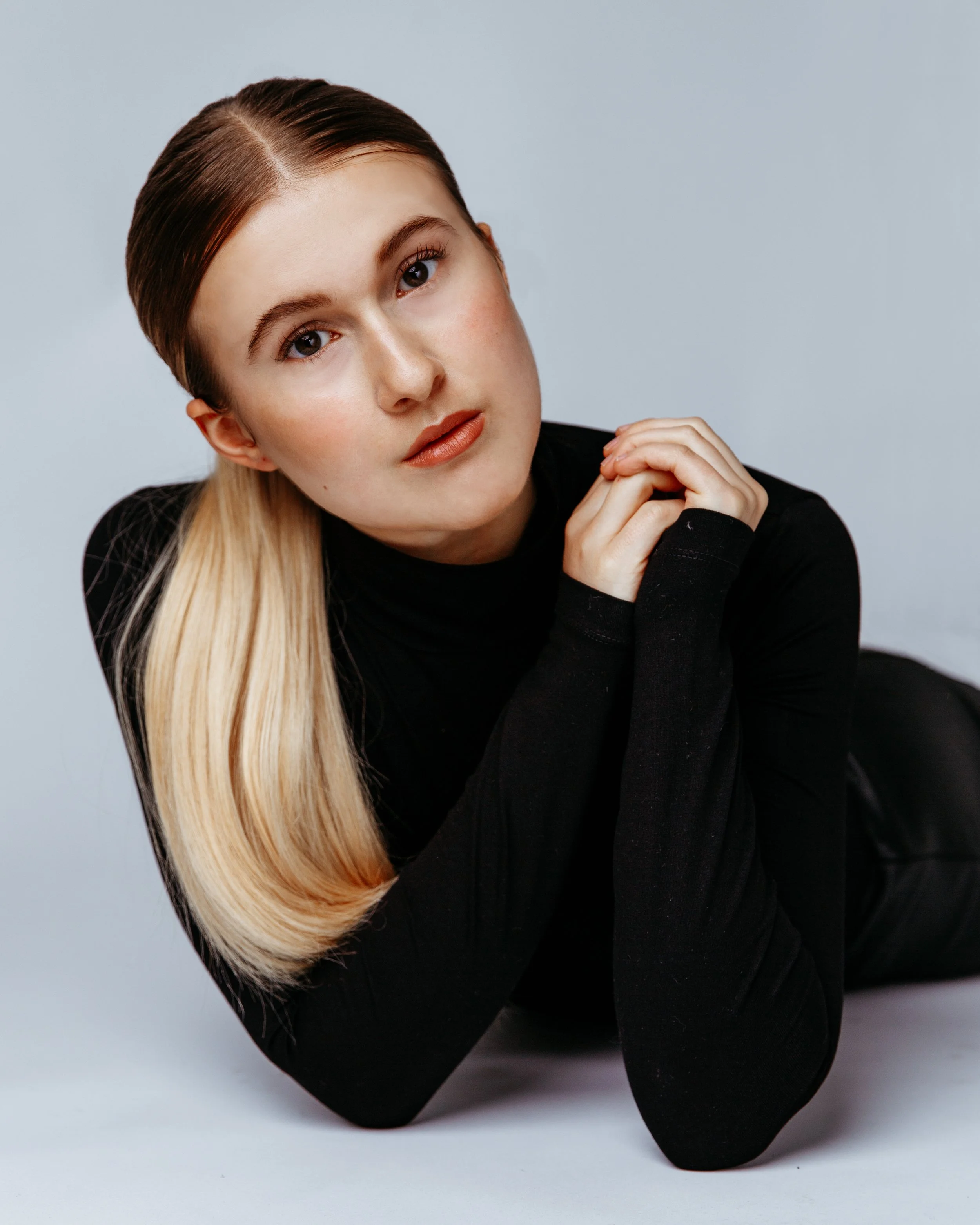 A woman with blonde hair, styled straight, wearing a black top, looking directly at the camera with her chin resting on her hand against a gray background.