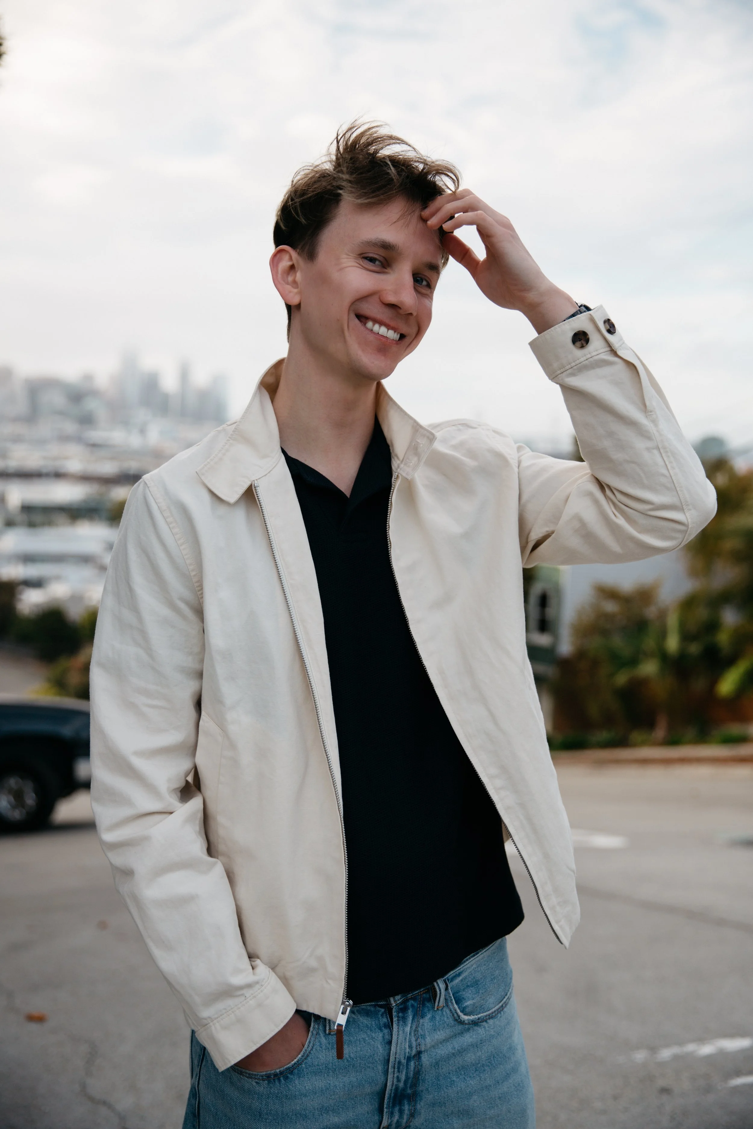 A young man with brown hair smiling and touching his head with his right hand, wearing a beige jacket, black shirt, and blue jeans, standing outdoors with a cityscape background and cloudy sky.