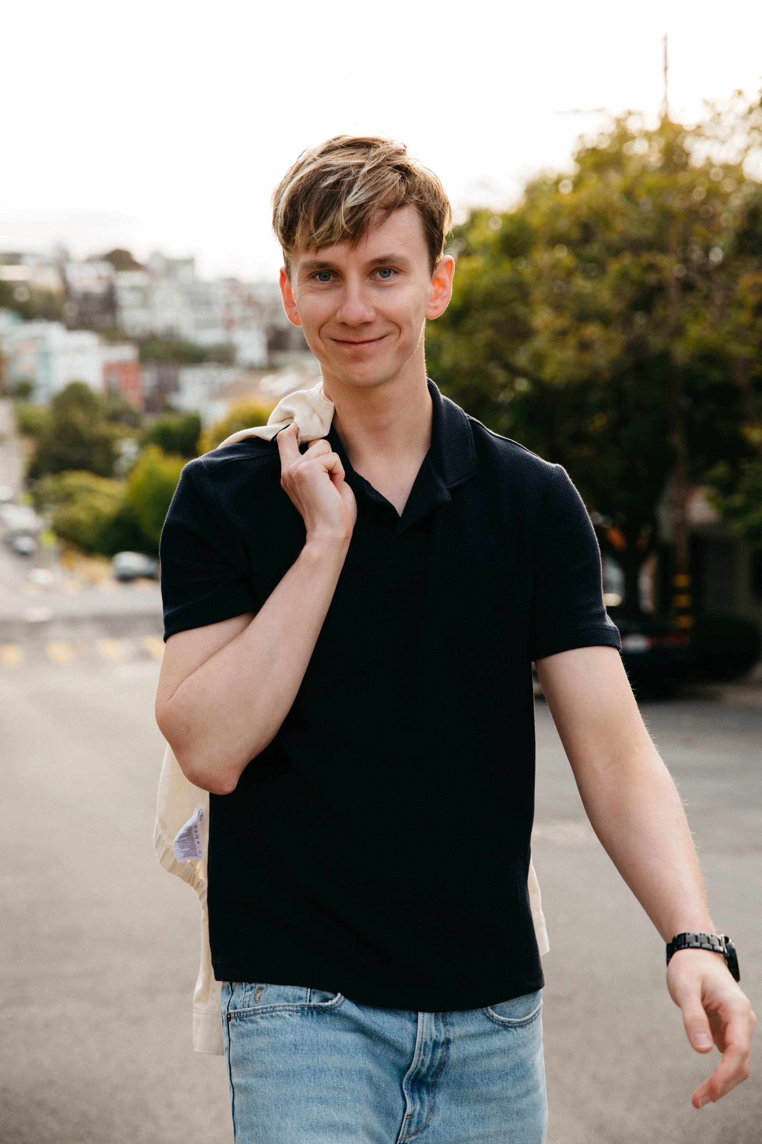 A young man with light brown hair and blue eyes smiling outdoors, wearing a black polo shirt, light blue jeans, and a wristwatch, with a beige jacket over his shoulder, in front of blurred cityscape and green trees.