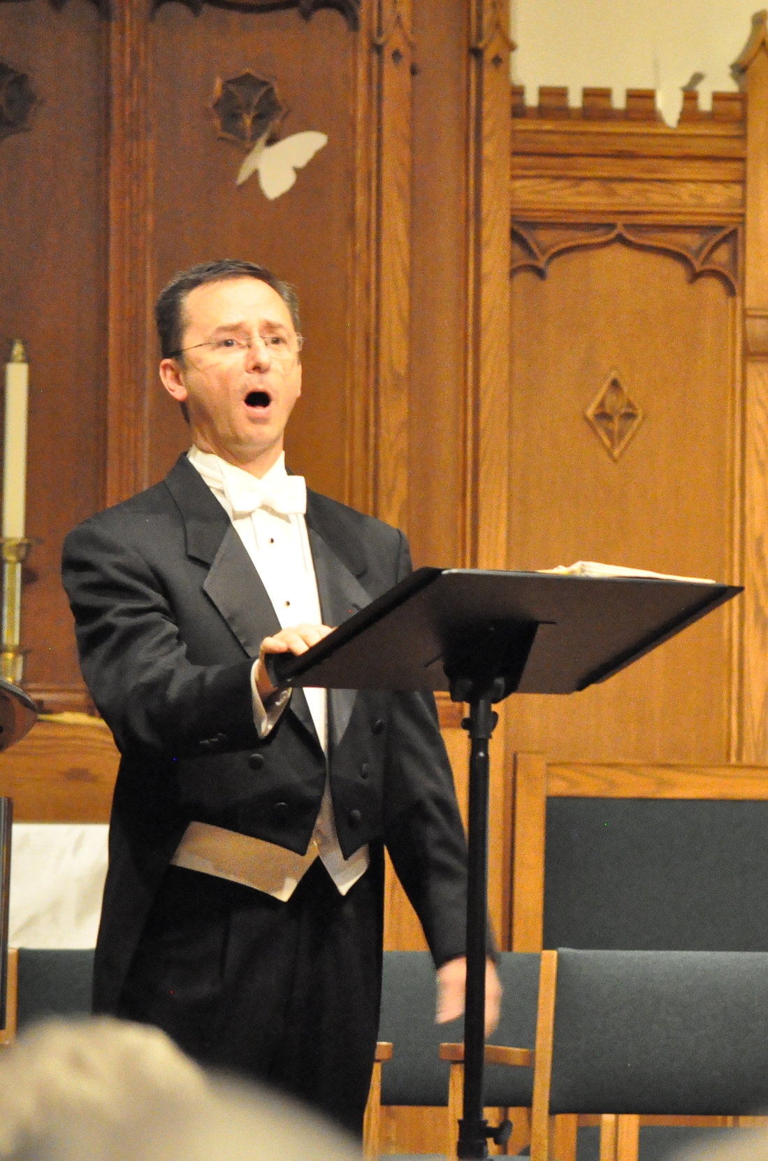 Chuck Taft  performing as a choir conductor or soloist, standing at a music stand in a church or concert hall with wooden paneling and carved details on the wall.