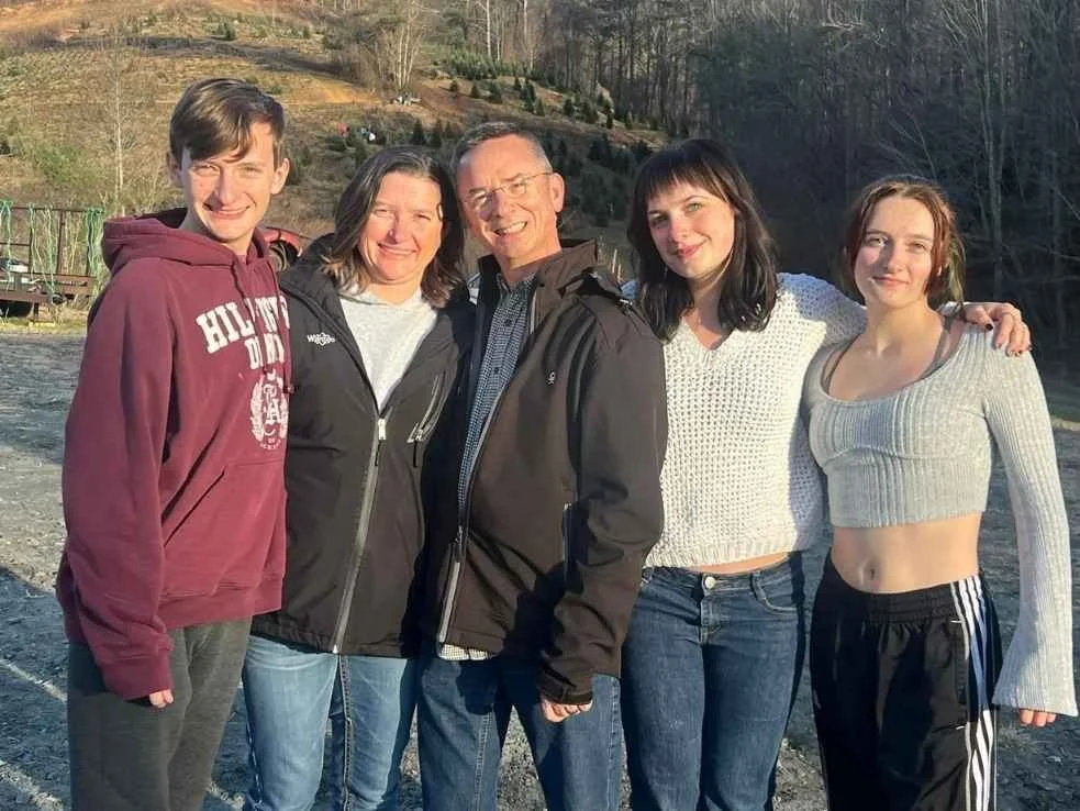 Chuck and Renee standing outdoors in a line, smiling, with hills and trees in the background with their three children.