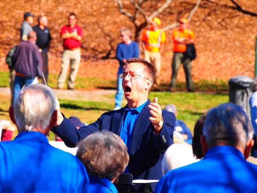 A man in a blue shirt and dark blazer is speaking passionately to an audience outdoors, with several people in the background and some audience members in blue jackets facing him.