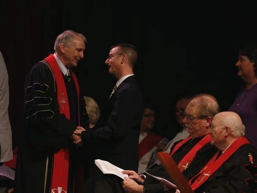 A graduation ceremony with individuals in academic regalia shaking hands and receiving diplomas.