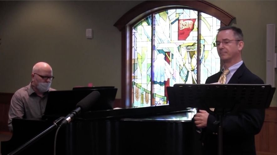 A man in a suit and tie singing or speaking at a music stand, next to a pianist wearing glasses and a face mask, in front of a decorated stained glass window with religious motifs in a church or chapel.