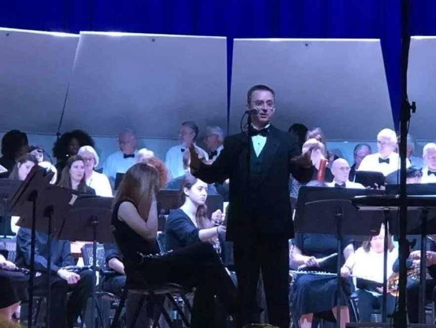 Conductor in a tuxedo standing on stage before a large orchestra performing in a concert hall
