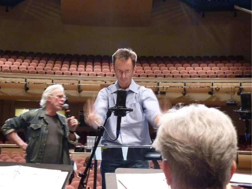 A rehearsal or recording session in an empty theater or concert hall, with two men at the front, one with a microphone and the other adjusting equipment, and a person with gray hair and glasses in the foreground.