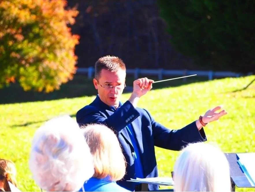 A man conducting an outdoor choir or orchestra on a sunny day, with a group of older women listening.