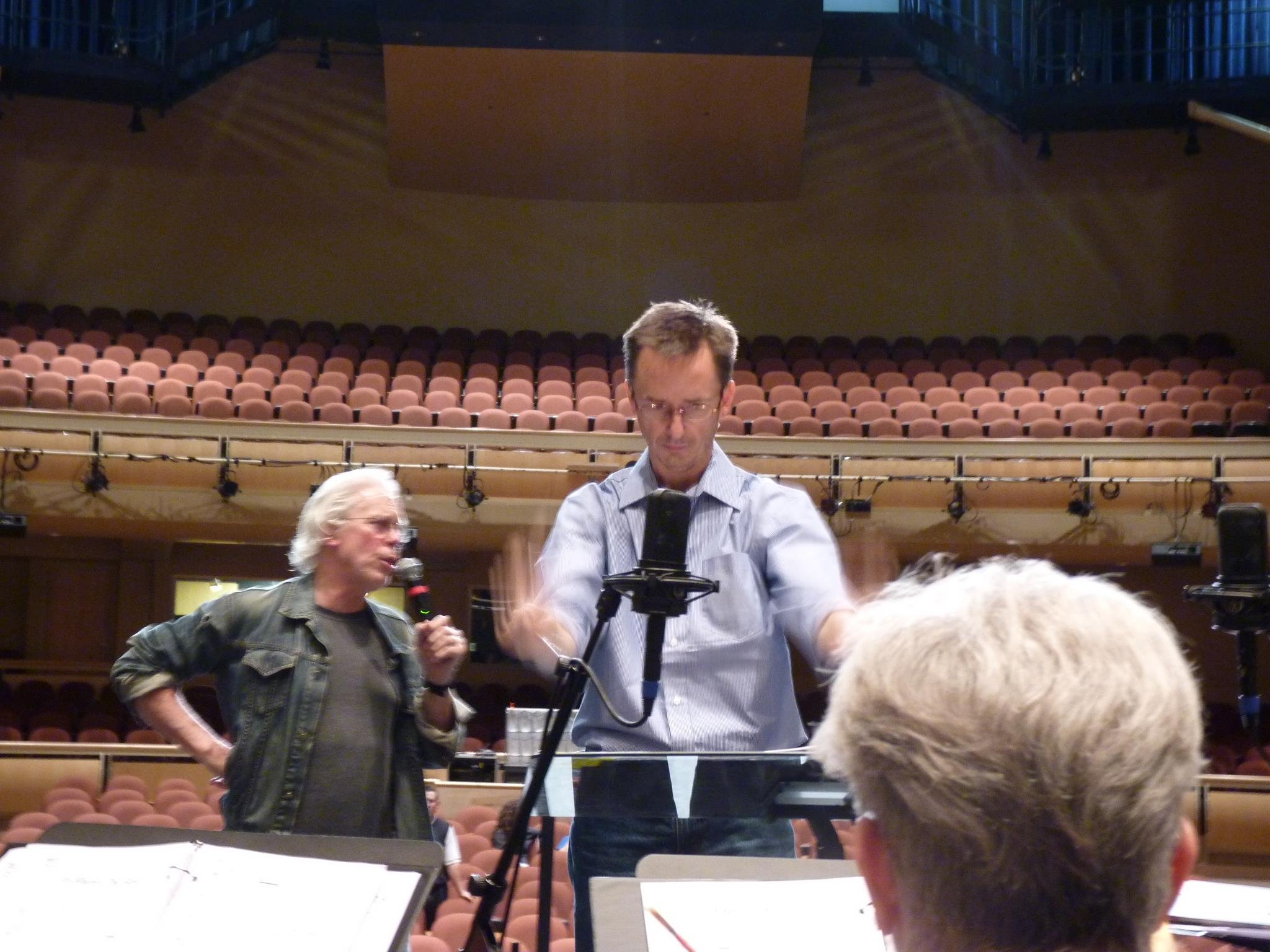 A rehearsal or recording session in an empty theater or concert hall, with two men at the front, one with a microphone and the other adjusting equipment, and a person with gray hair and glasses in the foreground.