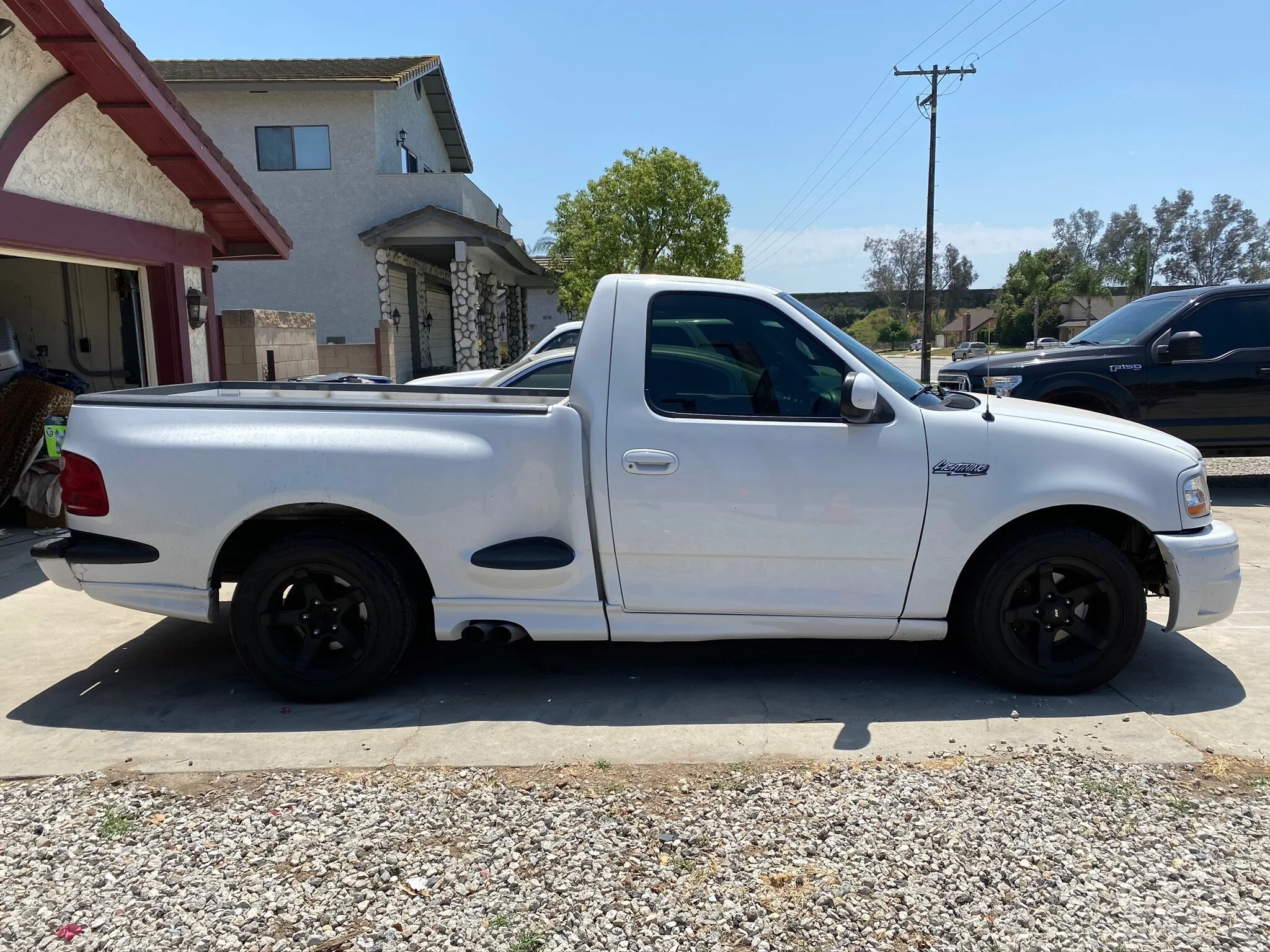 White pickup truck parked in driveway of residential neighborhood, with several other vehicles visible and houses in the background.