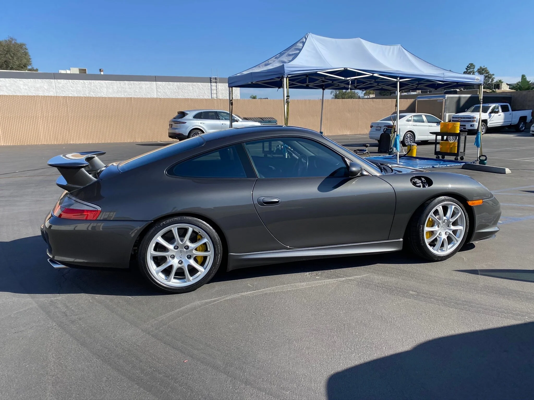 Gray Porsche sports car parked in a lot near a tent with maintenance supplies, with other vehicles in the background.