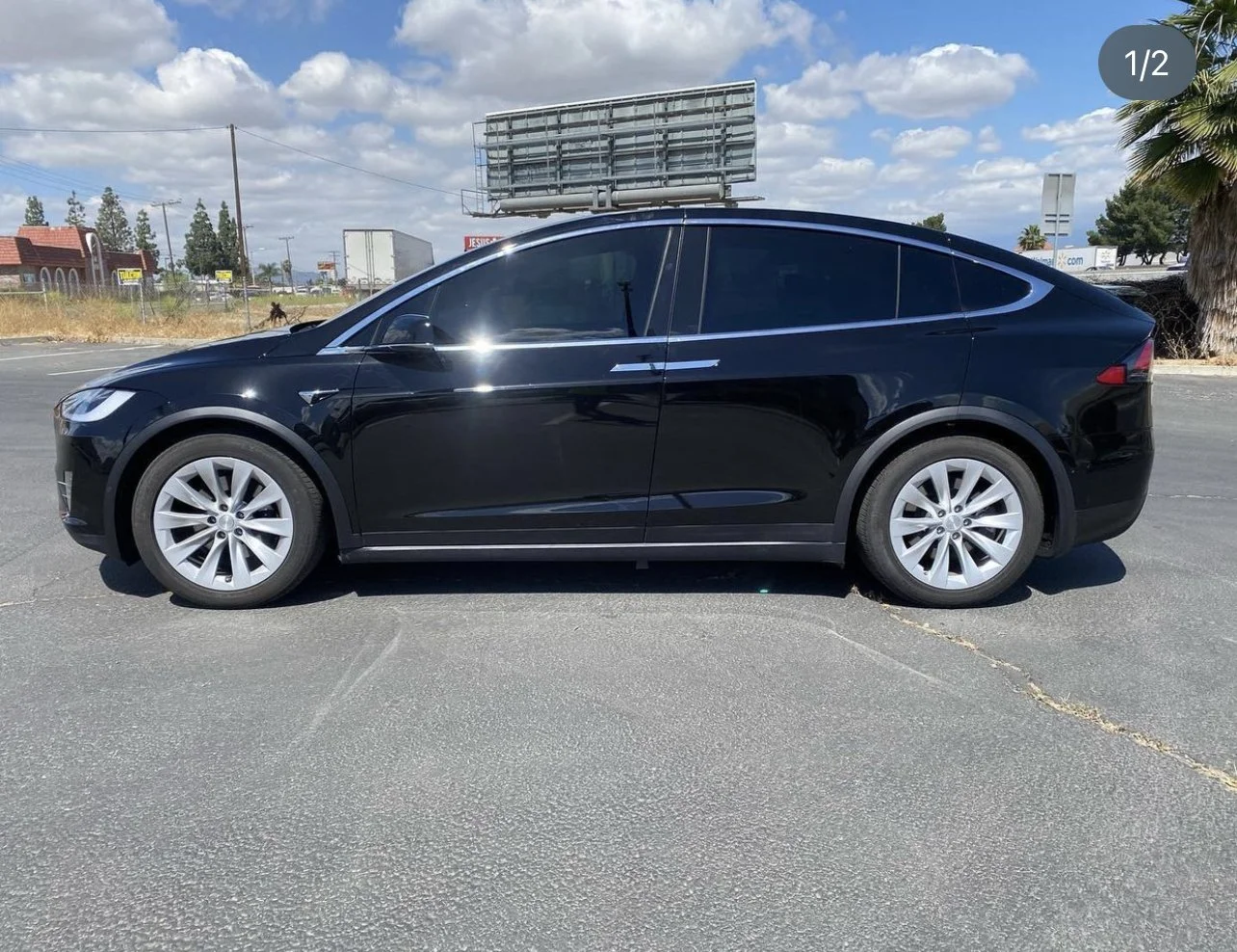 Black Tesla Model X parked on an asphalt lot under partly cloudy sky with a billboard and palm trees in the background.