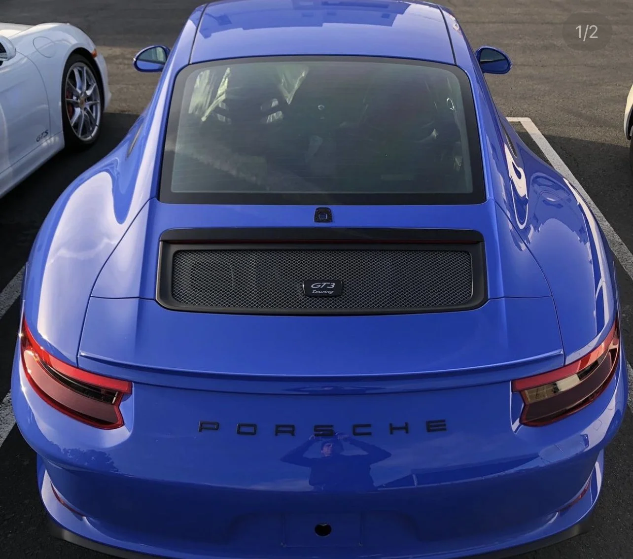 Back view of a blue Porsche GT3 Touring parked in a parking lot, with the Porsche logo and a rear light visible.