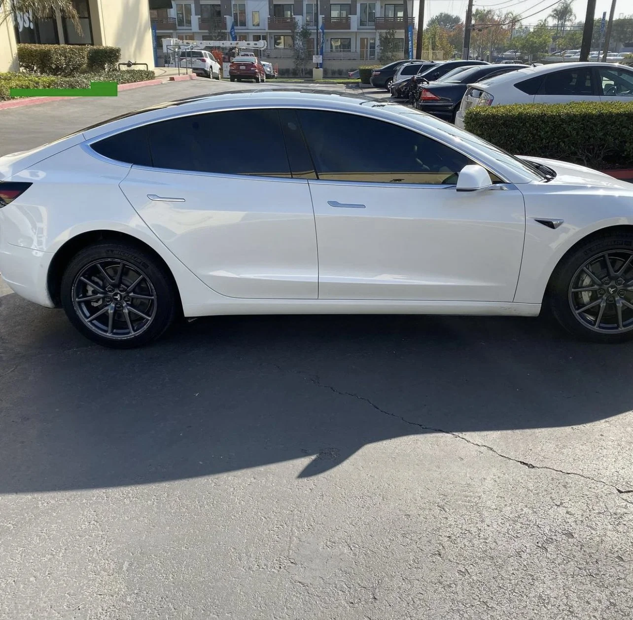 White Tesla Model 3 parked in a parking lot with other cars visible in the background and a building behind.